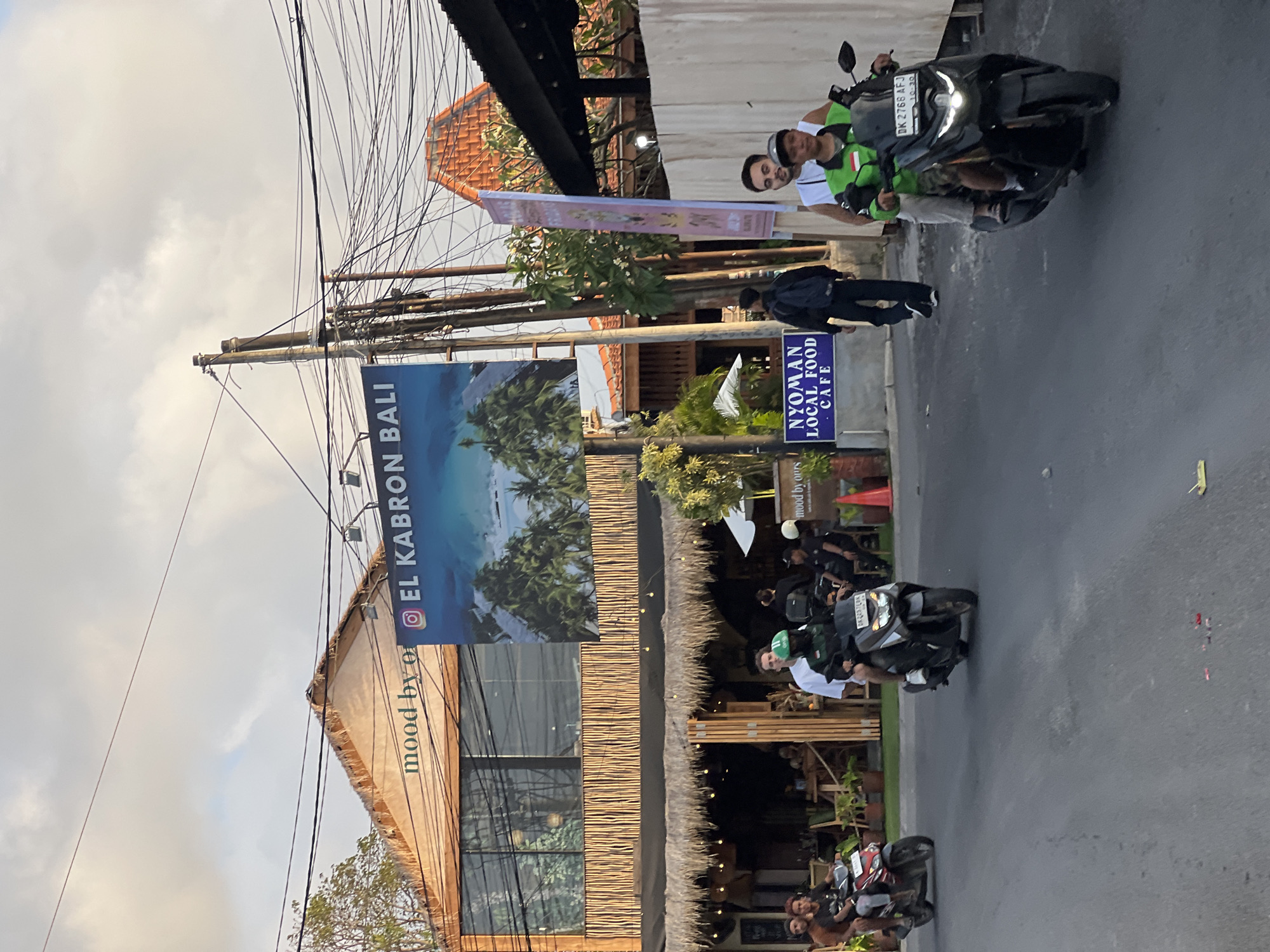 In Kuta Selatan, Indonesia, a street scene unfolds under a cloudy sky with power lines crisscrossing overhead. A building with a thatched roof and wooden exterior, identified by the sign "mood by ours" and an Instagram logo alongside "EL KABRON BALI," dominates the background. Below, a cafe with an open-air seating area is visible, with patrons seated at tables. Two motorbikes are prominent in the foreground: one red ridden by two individuals and another black one carrying two people, one in a green vest. A third black motorbike, ridden by a person in a green vest, is closer to the viewer, with a visible license plate "DK 2766 AFJ." To the left of the building, a blue sign reads "NYOMAN LOCAL FOOD CAFE." The street is paved, and the overall atmosphere suggests a casual, tropical setting.
