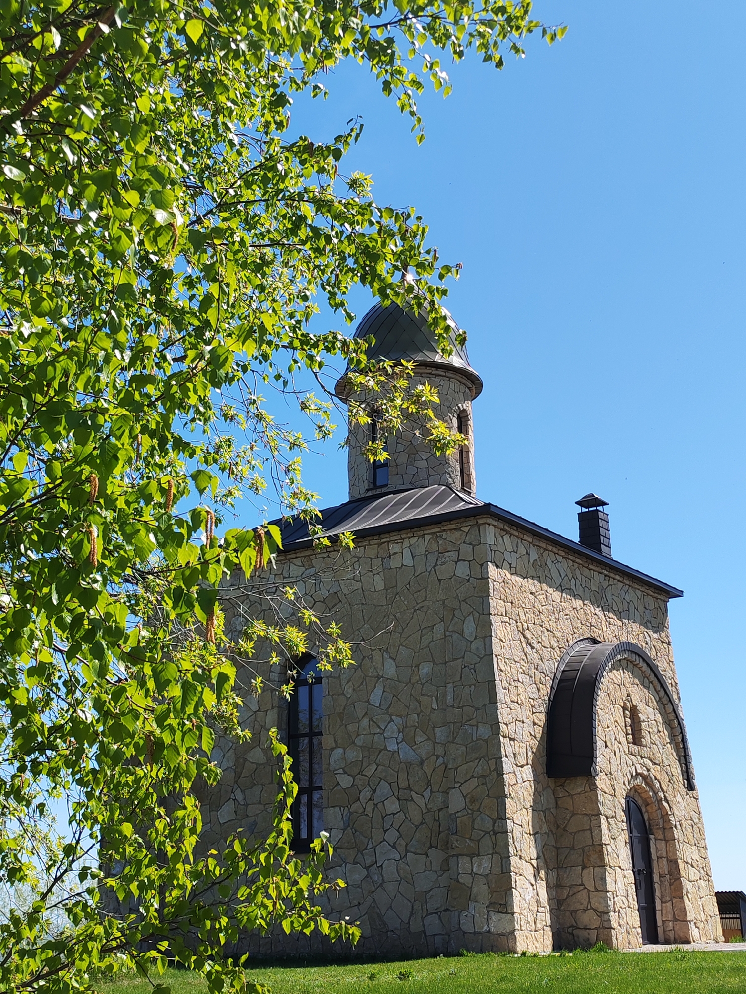 A ground-level, medium shot captures a stone building with an attached tower and a background of clear, blue sky. The building, constructed of light tan stones, has a dark metal roof and a chimney. The tower has a dark metal dome. A tree with bright green leaves is visible in the left side of the frame, partially obscuring the building. The building sits on a patch of green grass. The location is likely Икское Устье, Russia. No people are visible in the frame.