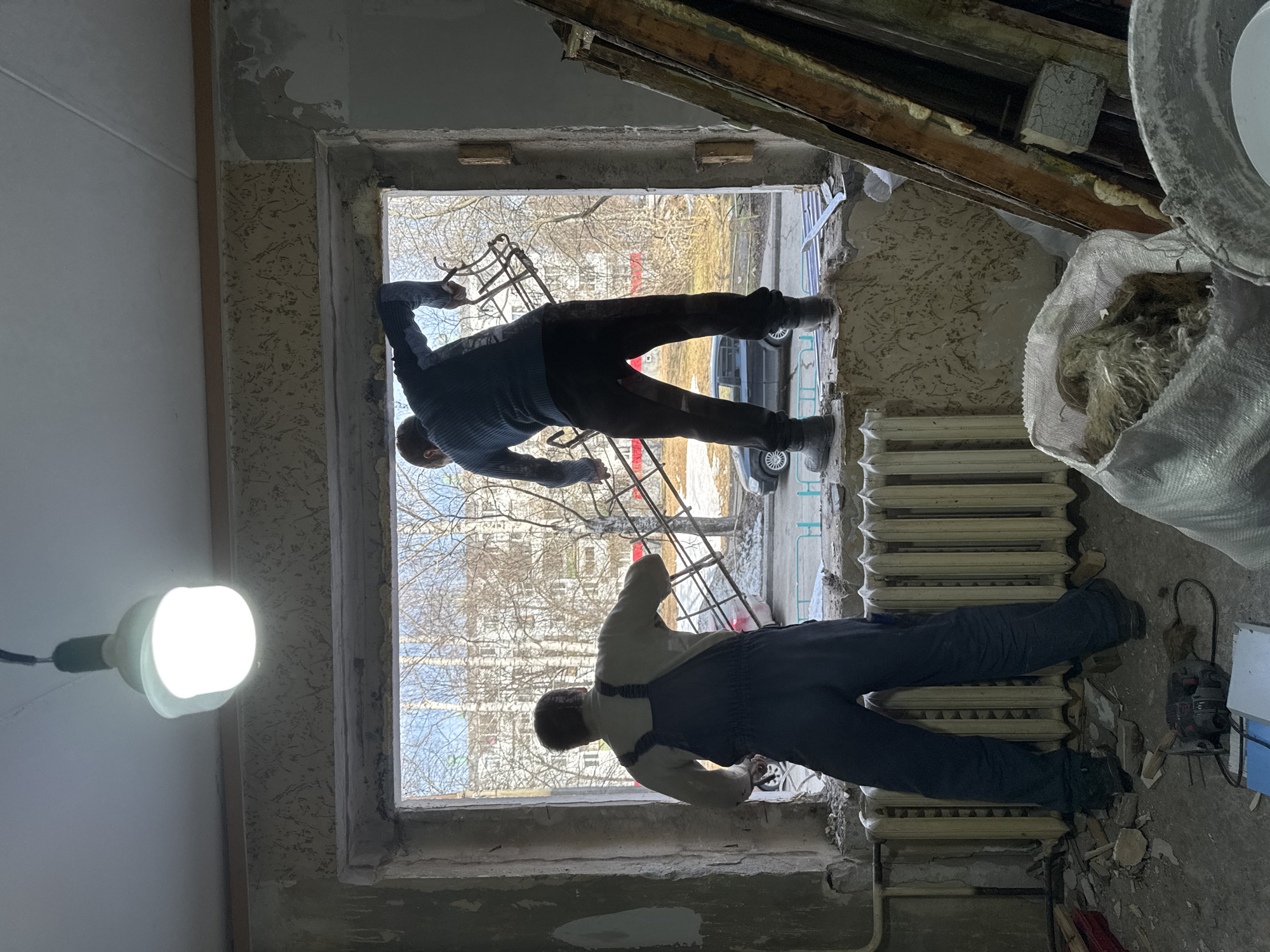 Two workers are installing a window in an apartment renovation in Chaikovsky, Russia. The window frame is open to the outside view of a street with buildings, trees, and a parked car. The workers are positioned on opposite sides of the window. One is inside, wearing blue overalls and the other is partially outside, wearing a blue sweater. Debris and construction materials are scattered around the room. A light bulb hangs from the ceiling. A white radiator is located below the window to the right.