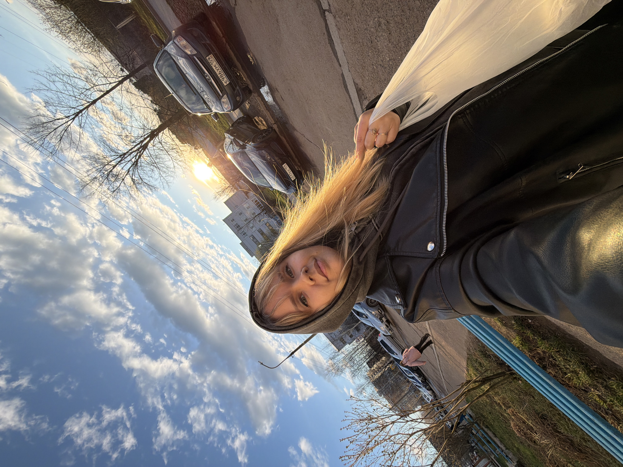 This outdoor image is a tilted, wide-angle shot, likely a selfie, capturing a young woman and her surroundings during the golden hour. The main subject is a blonde woman wearing a dark hooded jacket, looking directly at the camera with a neutral expression. A white plastic bag is draped over her left shoulder, and a ring is visible on her right hand.
The background features a street scene with parked cars, leafless trees, and multi-story buildings under a bright sky with scattered white clouds. The sun is low, casting a warm, golden glow across the scene, typical of either sunrise or sunset. Several cars are parked along a paved road. Visible license plates on two cars, "E986XM 152" and "051TEE 152," indicate a Russian location (Nizhny Novgorod Oblast). A blue, striped structure, possibly a fence or utility pipes, runs vertically along the right edge of the frame. Power lines are visible against the sky. The setting suggests an urban or suburban environment in late winter or early spring.