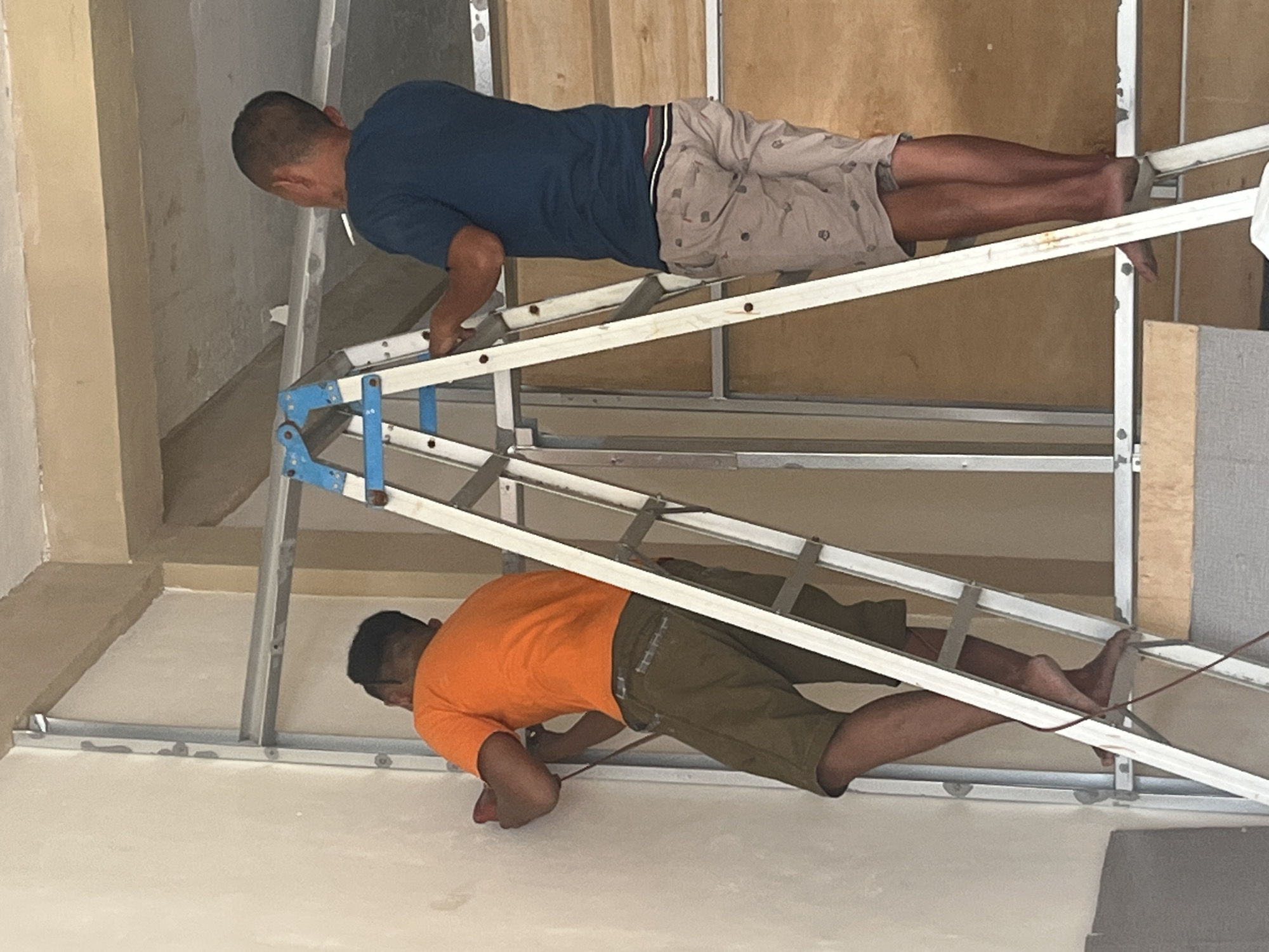 Two men are working on a construction site in Sekayu, Indonesia. The man on the left, wearing an orange shirt and brown shorts, is standing on a ladder and using a red power tool on a white wall. The man on the right, wearing a dark blue shirt and patterned shorts, is also on a ladder, holding a cigarette. The site appears to be indoors, with metal framing and wooden panels visible. The overall scene suggests ongoing construction or renovation work.