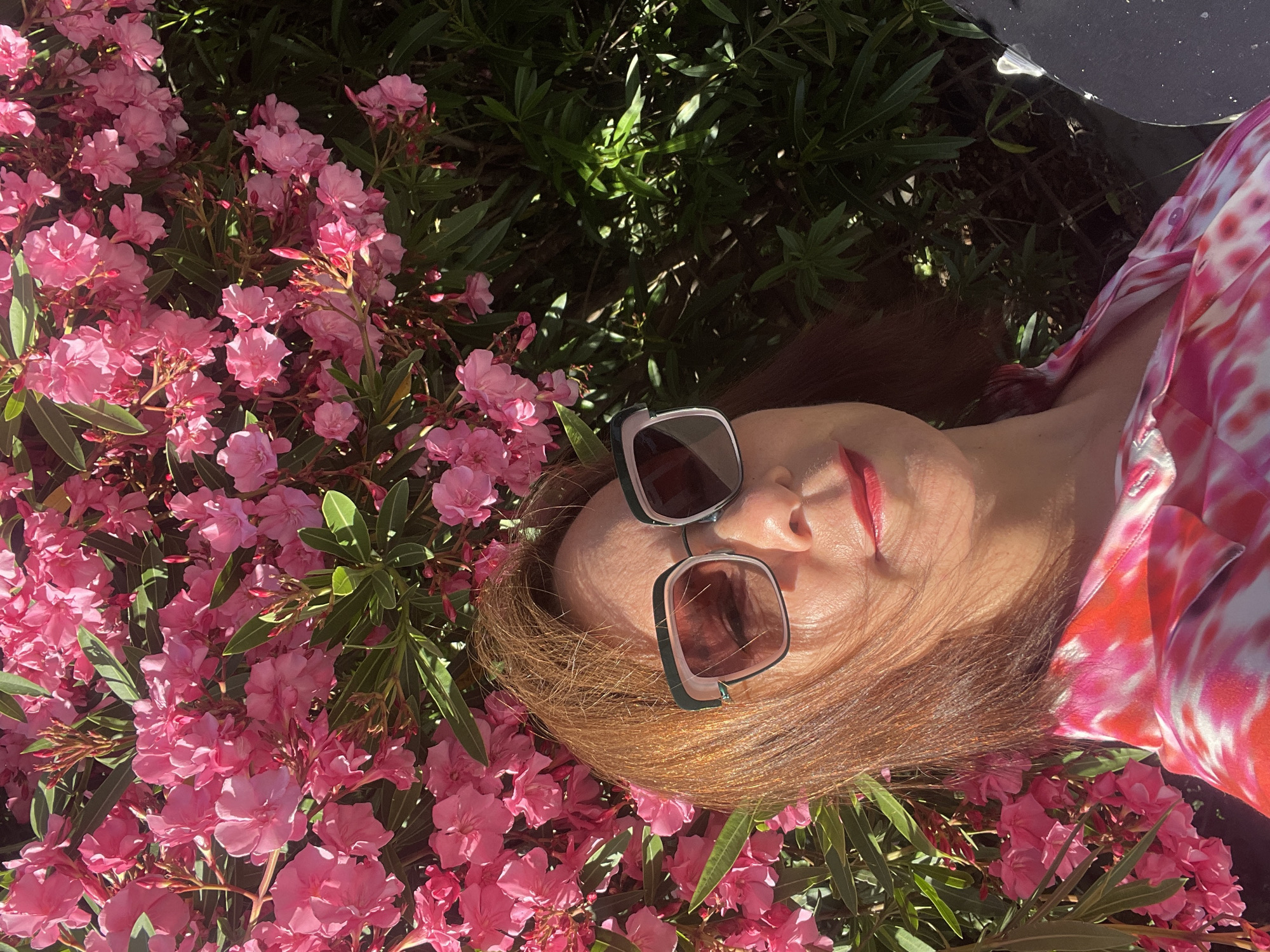 A woman with reddish-brown hair and square-shaped sunglasses is taking a selfie in front of a large bush of pink oleander flowers. She is wearing a pink and white tie-dye shirt. The image is taken outdoors in daylight, with dappled sunlight filtering through the leaves and flowers. The background consists of lush green foliage and the vibrant pink blooms of the oleander. The specific location in Sochi, Russia cannot be determined from the image alone.