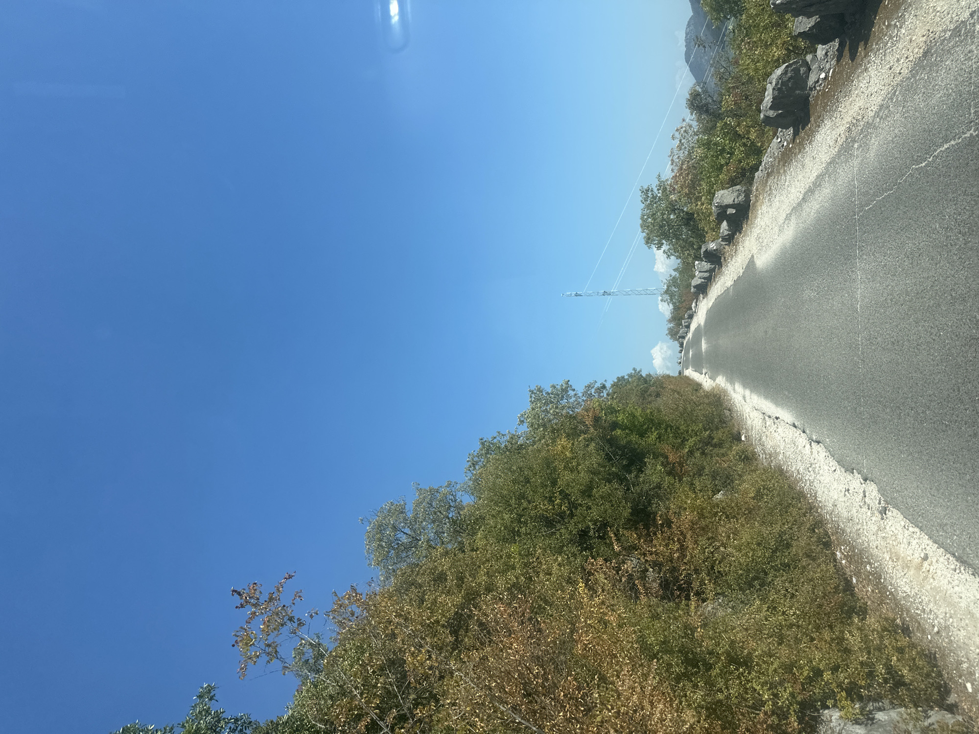 A paved road curves upward through a mountainous landscape under a clear blue sky. To the left, a steep, rocky embankment is covered in dense, green and brown foliage, with some trees showing autumnal colors. To the right, the road is bordered by a rough, gravelly shoulder and scattered large rocks. In the distance, a tall transmission tower stands on a hill, and beyond that, rolling hills fade into the horizon. The scene is devoid of people or vehicles. This image depicts a section of a road in the vicinity of Braćeni, Montenegro, characterized by its rugged terrain and natural vegetation.