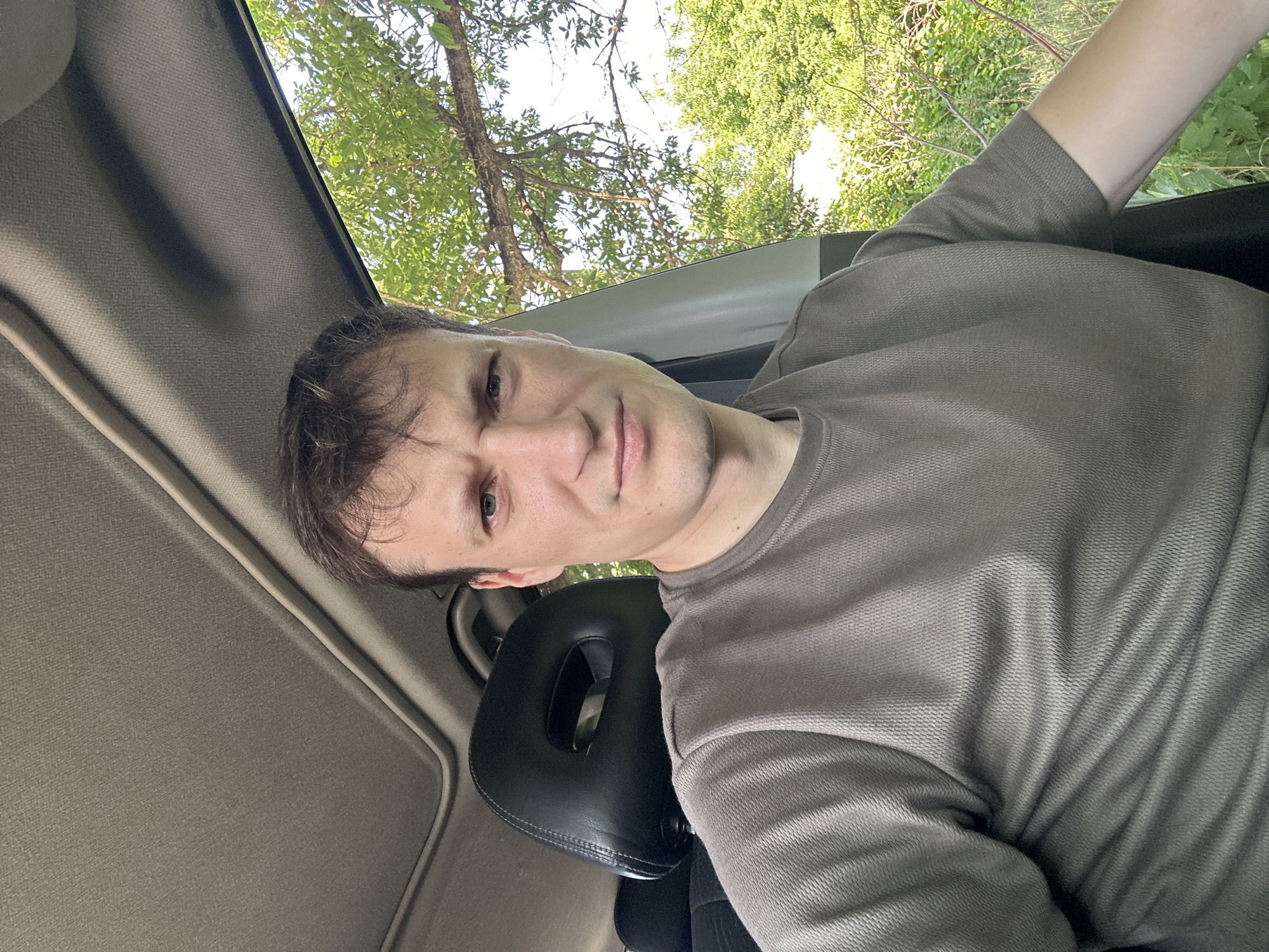 A man is sitting in the driver's seat of a car, looking at the camera. The car's sunroof is open, and green trees are visible outside the open window. The man is wearing a brown t-shirt. The photo was taken in Makiv, Ukraine.