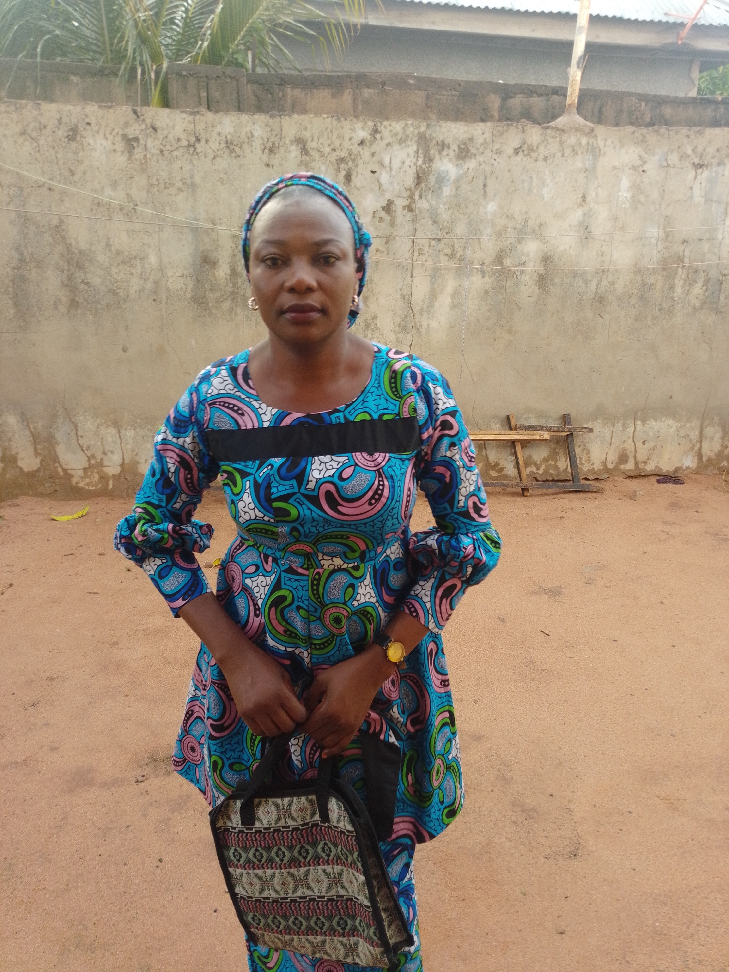 A woman stands outdoors in front of a concrete wall. She is wearing a colorful, patterned blue dress with pink and green accents, and a matching headscarf. She is holding a patterned bag with black handles. A gold watch is visible on her left wrist. The ground is sandy and there is some debris scattered around, including a yellow leaf and a small wooden structure in the background. The wall appears weathered and the sky is not visible.