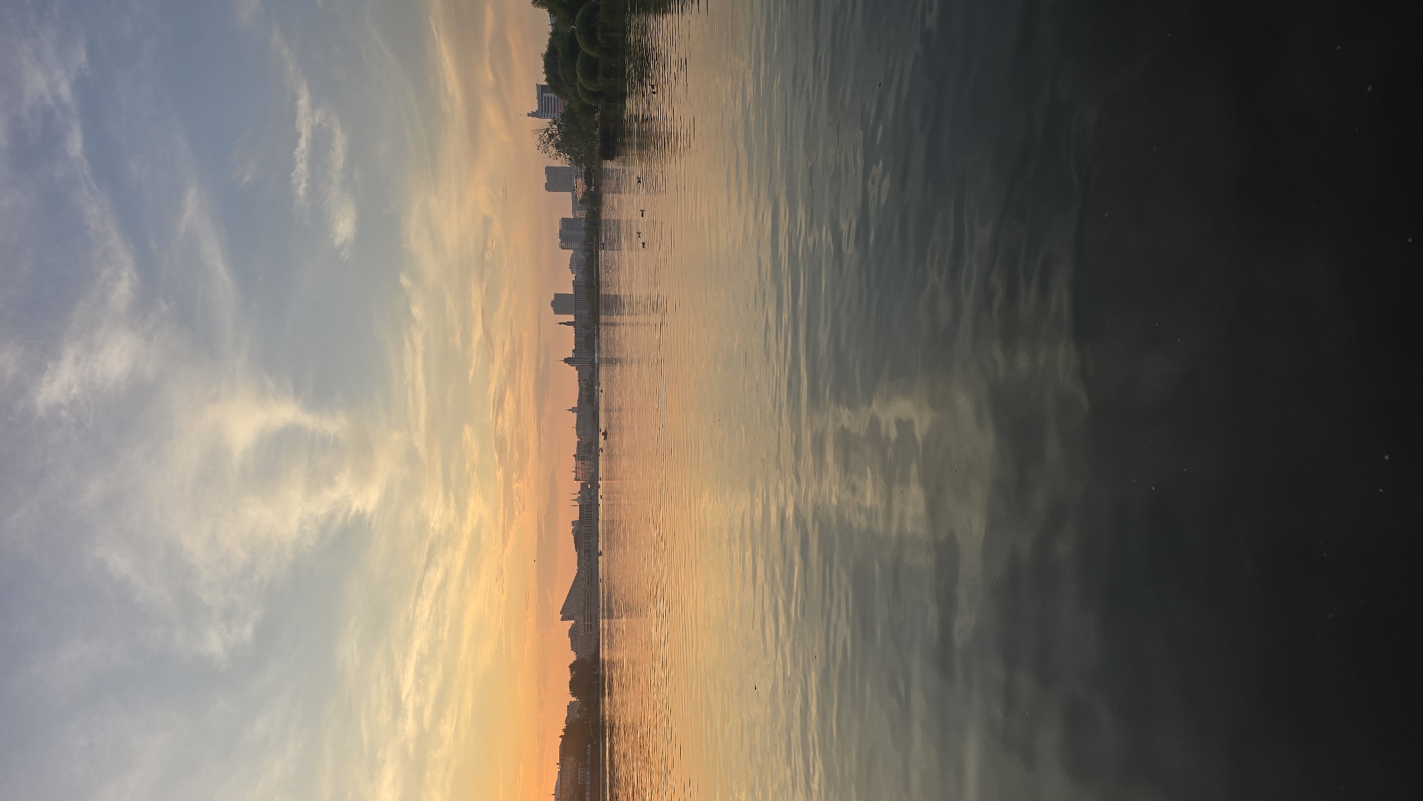 The image depicts a serene lakeside scene in Kazan, Russia, at sunset. The sky is a gradient of warm orange and soft yellow near the horizon, transitioning into a pale blue higher up, with wisps of white clouds scattered throughout. In the background, the Kazan skyline is visible, featuring a collection of buildings, including tall modern structures and older, more traditional architecture. The city lights are beginning to twinkle, hinting at the approaching night.
The lake in the foreground is calm, with gentle ripples reflecting the colors of the sunset and the city lights. Several ducks are scattered across the water's surface. A few small boats can be seen further out on the lake, suggesting recreational activity. The overall atmosphere is tranquil and picturesque, capturing a moment of natural beauty within an urban setting.