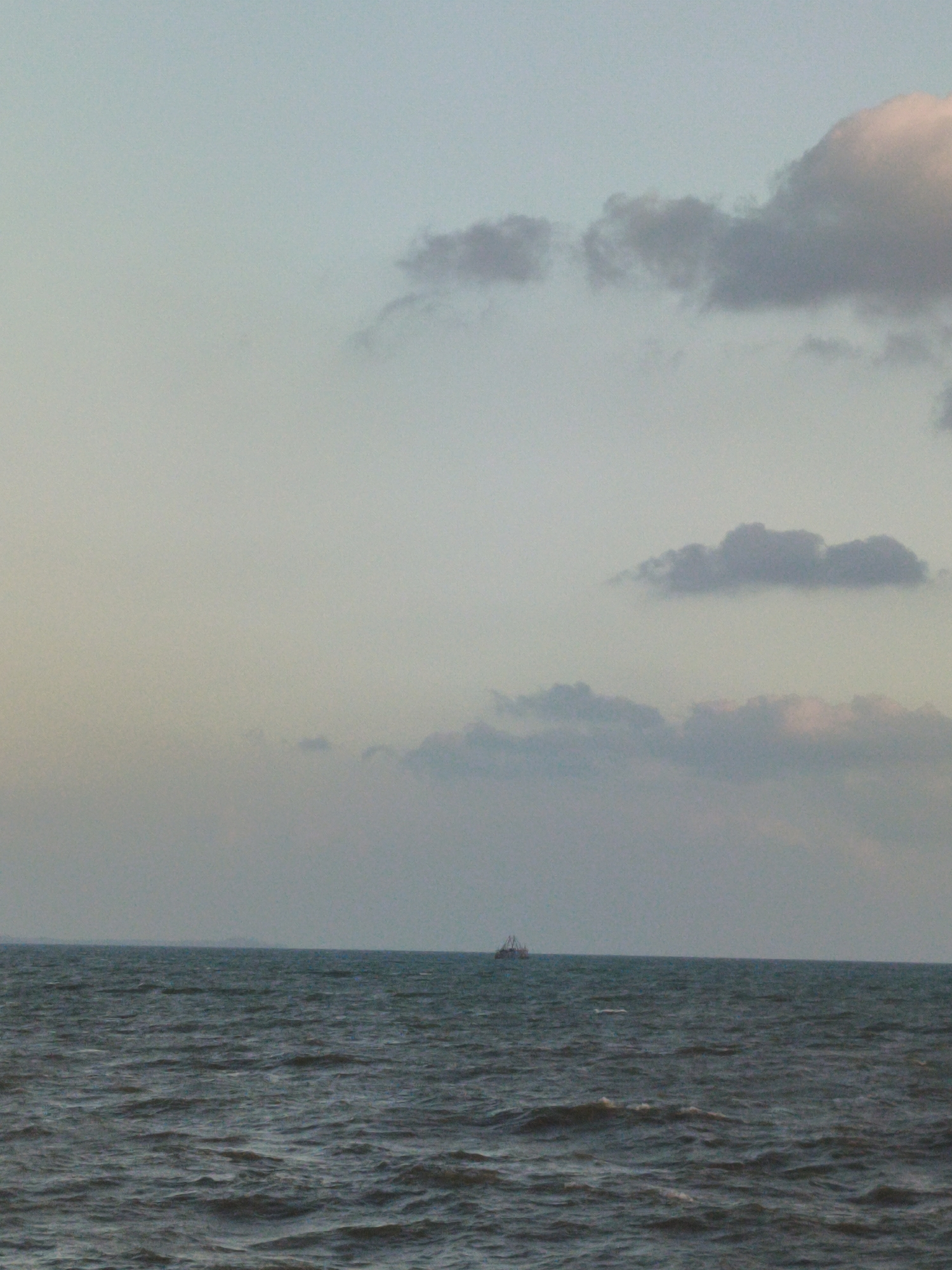 A distant boat is visible on the horizon of a body of water. The sky above is a pale blue with scattered clouds of various shades of gray and pink. The water appears dark and choppy, with small waves. The scene is set during what appears to be either dawn or dusk, given the soft lighting and muted colors. The location is the city of Kupang Berkah Jaya, Indonesia.