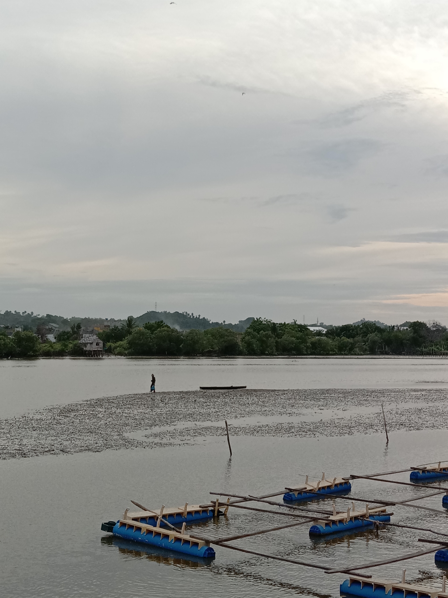 The image depicts a coastal landscape in Lhokseumawe, Indonesia, under an overcast sky. In the foreground, several blue plastic floatation devices are arranged in rows on the water's surface, connected by wooden beams. In the middle ground, a muddy sandbank stretches across the water, with a solitary figure walking on it. Further along the sandbank, a small boat rests. The background features a shoreline with trees, houses, and a distant hill topped with communication towers.
