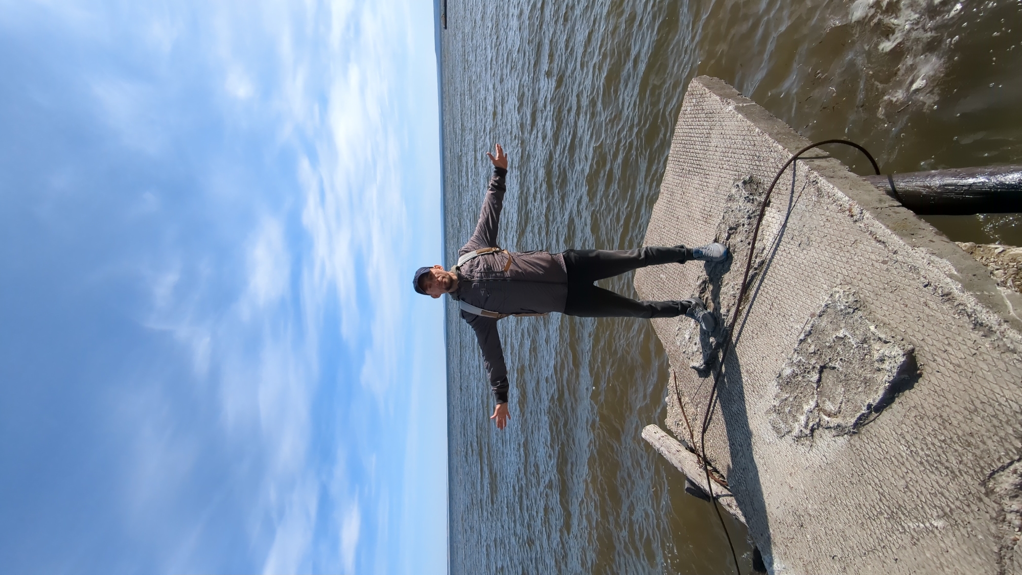 A man stands with outstretched arms on a weathered concrete structure next to a large body of water under a bright sky. The image, taken during the daytime, shows the man centered, facing away from the camera and towards the expanse of water, his posture suggesting balance or a feeling of freedom. He is wearing a dark jacket, dark pants, and a dark baseball cap, with what appears to be straps or a harness visible over his jacket. The concrete he stands on is rough and textured, possibly part of a pier or breakwater, and shows signs of wear, with some exposed material resembling rebar or mesh. A wooden log or pole is partially visible near the concrete structure on the right. The water is dark and choppy, extending to a clear, distant horizon. The sky above is a vibrant blue with scattered white, wispy clouds. No visible text is present.