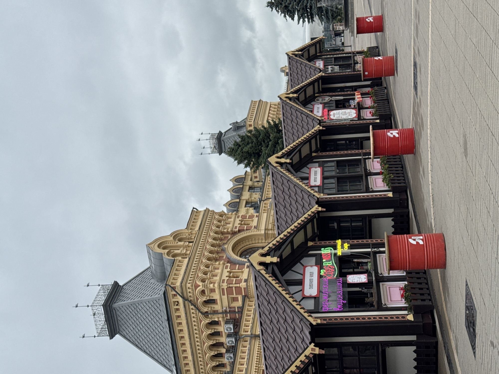 The image depicts a cloudy day scene in Nizhny Novgorod, Russia, specifically on Bolshaya Pokrovskaya Street. In the foreground, a row of small, dark brown market stalls with brown roofs and ornate yellow trim is visible. These stalls are arranged side-by-side on a grey paved walkway. Each stall has a counter where items appear to be sold, and some have neon signs in pink and green. One sign prominently reads "фермерские вкусняшки" (farmer's treats). Red barrels with a white symbol are placed in front of some of the stalls. Behind the stalls, a grand, ornate yellow and red building with a distinctive grey roof and a tower structure is visible, characteristic of historical architecture in Nizhny Novgorod. The sky is overcast with grey clouds. To the right, evergreen trees are present. The overall scene suggests a market or a street fair environment.