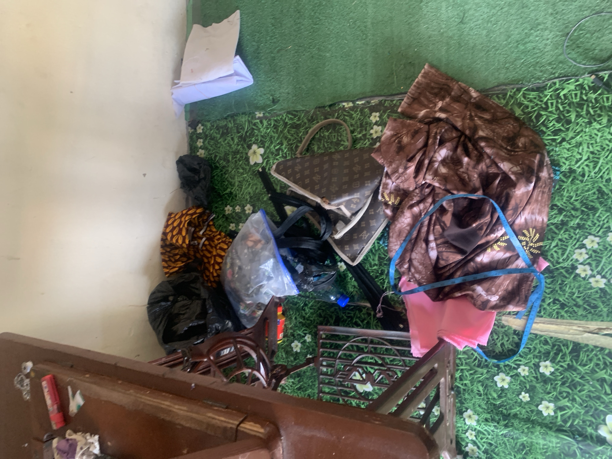 The image shows a cluttered indoor scene, likely a room in Jauro Yinu, Nigeria. A wooden table, possibly part of an old sewing machine, is on the left. On the table are various items including a red and silver lighter, some fabric scraps, and a small metallic object. The floor is covered with a green rug featuring a grass and flower pattern.
On the floor, a black trash bag is visible, along with a folded piece of fabric with a yellow and orange geometric pattern. Near these items is a clear plastic bag containing more fabric, and a dark bag. A brown designer handbag with a distinctive pattern is also present. Further to the right, there are folded white papers and a roll of green fabric. In the foreground, a pink fabric is laid out next to a brown patterned fabric, with a blue measuring tape draped over them. The overall impression is one of disarray and a possible craft or sewing area.