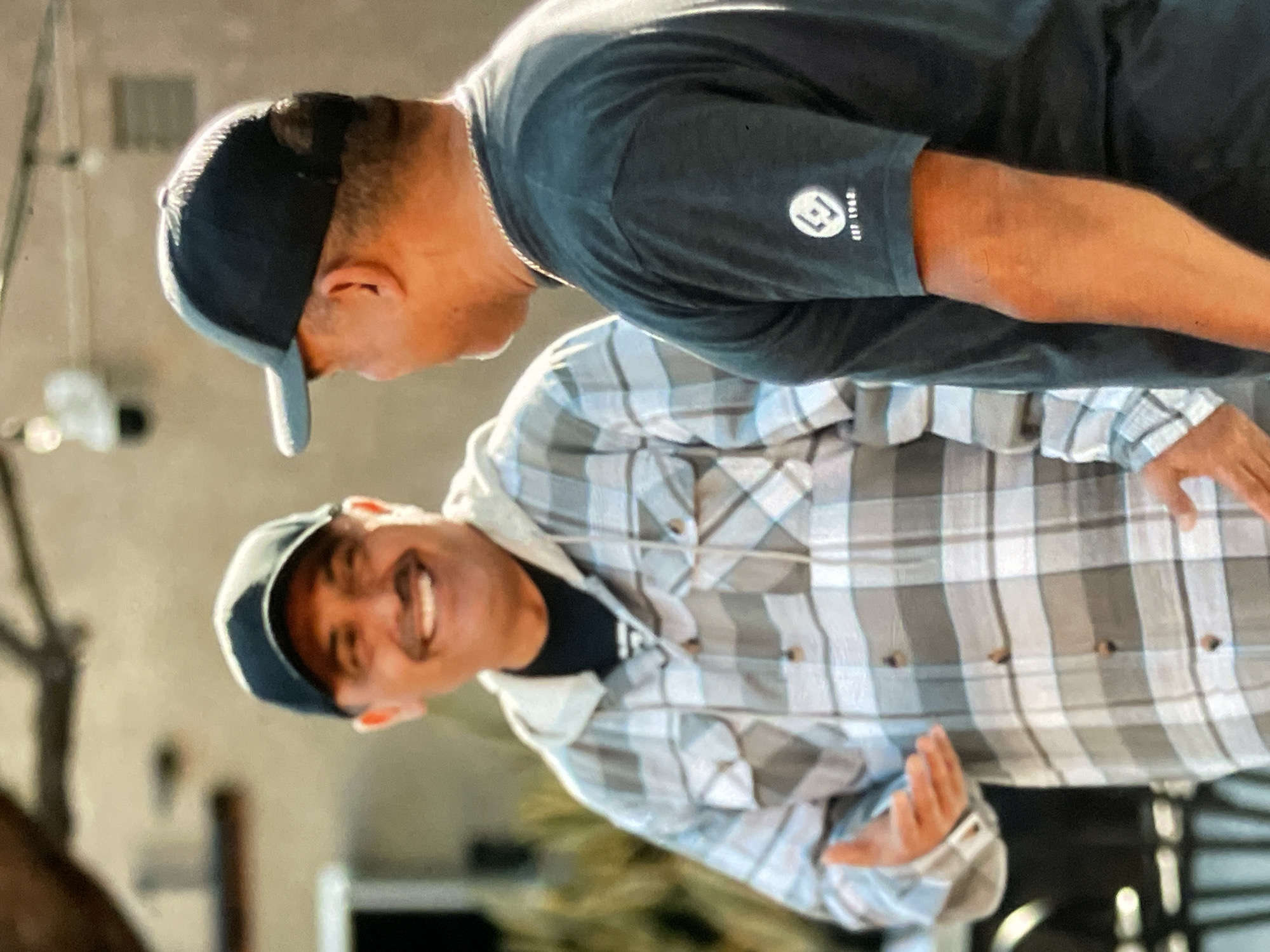 Two men are standing outdoors, facing each other. The man on the left is wearing a plaid shirt and a black baseball cap. He has a mustache and is smiling. His left hand is raised, palm open, in a gesture of speaking. The man on the right is wearing a dark t-shirt with a logo on the sleeve and a black baseball cap. He is turned slightly away from the camera, looking at the other man. The background suggests an urban environment, with buildings and some greenery visible. The lighting indicates it is daytime.