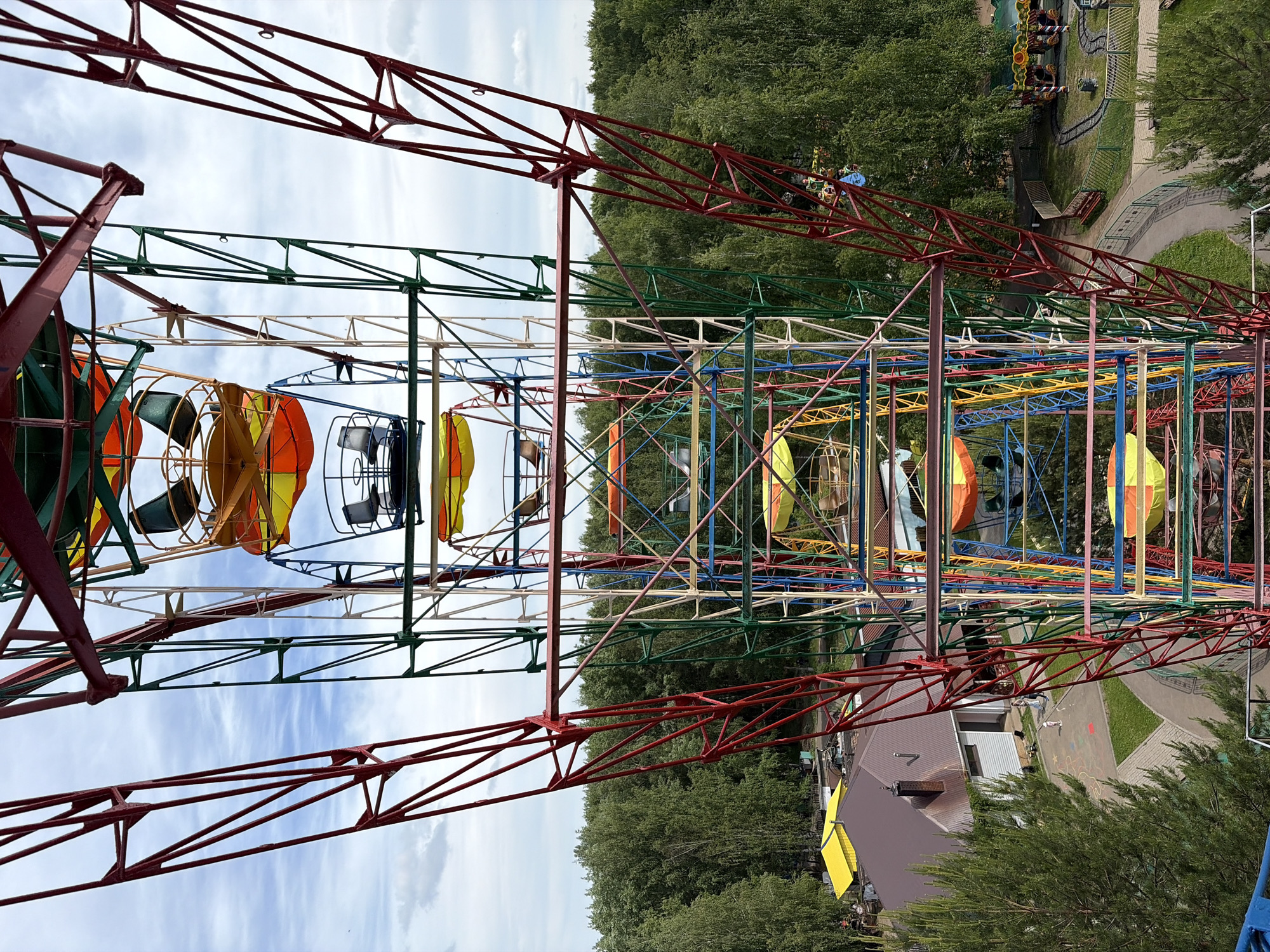 The image captures a vibrant amusement park scene from the perspective of someone on a large ride, likely a Ferris wheel, looking out and down. The foreground is dominated by the intricate metal framework of the ride, constructed from beams painted in a striking array of colors including red, green, blue, yellow, orange, and white. Several passenger cabins, some with colorful orange and yellow fabric coverings, and others in darker tones, are visible attached to the wheel's structure.
Beyond the ride's immediate framework, a sprawling landscape of dense green trees forms the backdrop, indicative of a large park or forested area. Below, sections of the amusement park become visible, featuring paved pathways, grassy areas, and what appears to be a miniature train track or roller coaster snaking through the foliage, with a small train visible on it. Several buildings with brown roofs and some with bright yellow awnings are nestled among the trees.
The sky is bright with scattered, wispy white clouds, indicating clear daytime weather. The overall impression is one of a cheerful and active outdoor environment. No discernible text is visible within the frame. This lively park setting is located in Chaikovsky, Russia.
