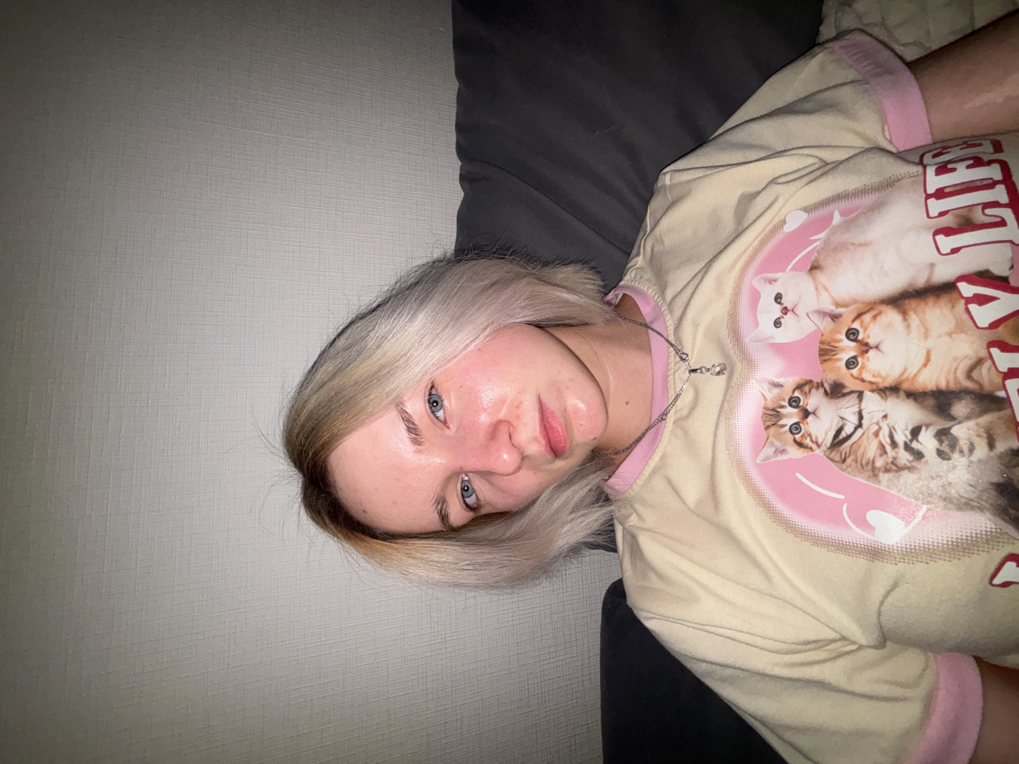 A person with blonde hair is lying on a dark gray couch against a light gray wall. They are wearing a beige shirt with pink trim and a graphic featuring multiple kittens. A silver chain necklace is visible. The location is Unknown, Russia.
