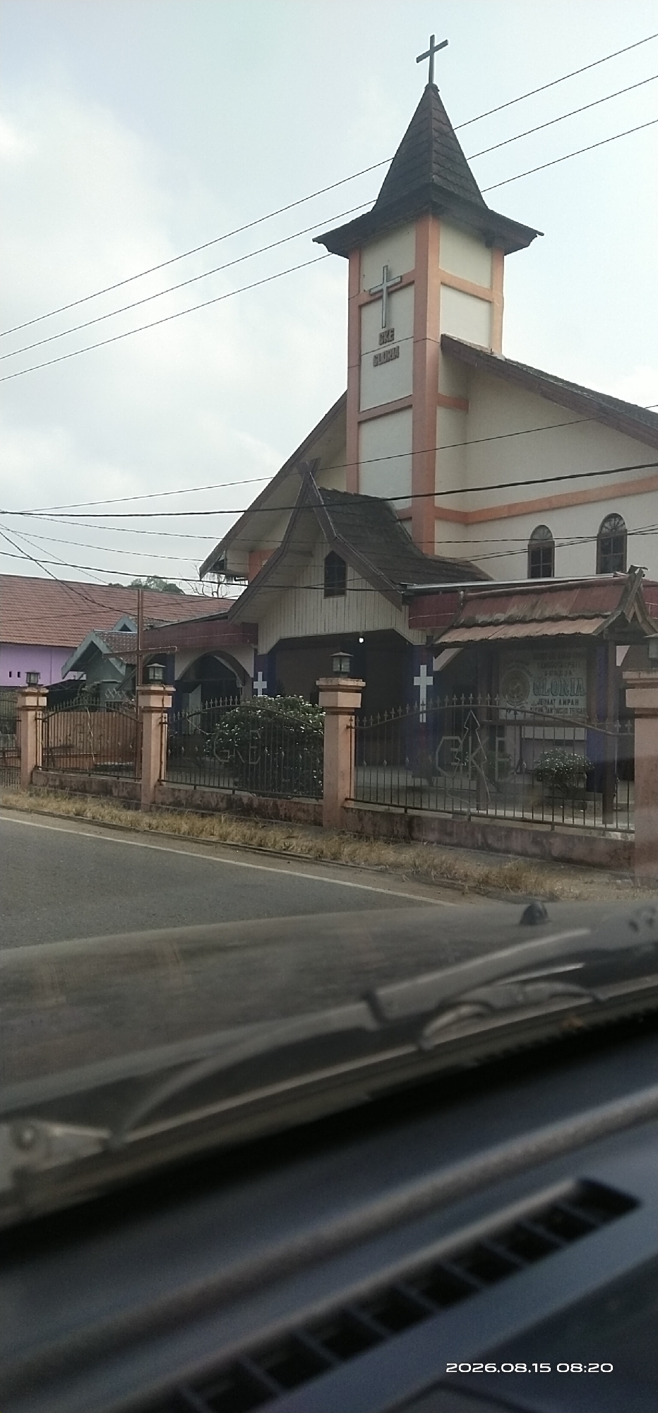 The image shows a church in Ampah Kota, Indonesia, with a prominent bell tower topped with a cross. The church building is white with peach-colored trim and features a pitched roof. A fence with decorative metalwork and pillars encloses the church grounds. The fence has crosses on it. A sign on the fence reads "GEREJA GLORIA." In the foreground, the dashboard and windshield of a vehicle are visible, suggesting the photo was taken from inside a car. The date stamp on the image indicates it was taken on August 15, 2026, at 08:20. The sky is overcast.