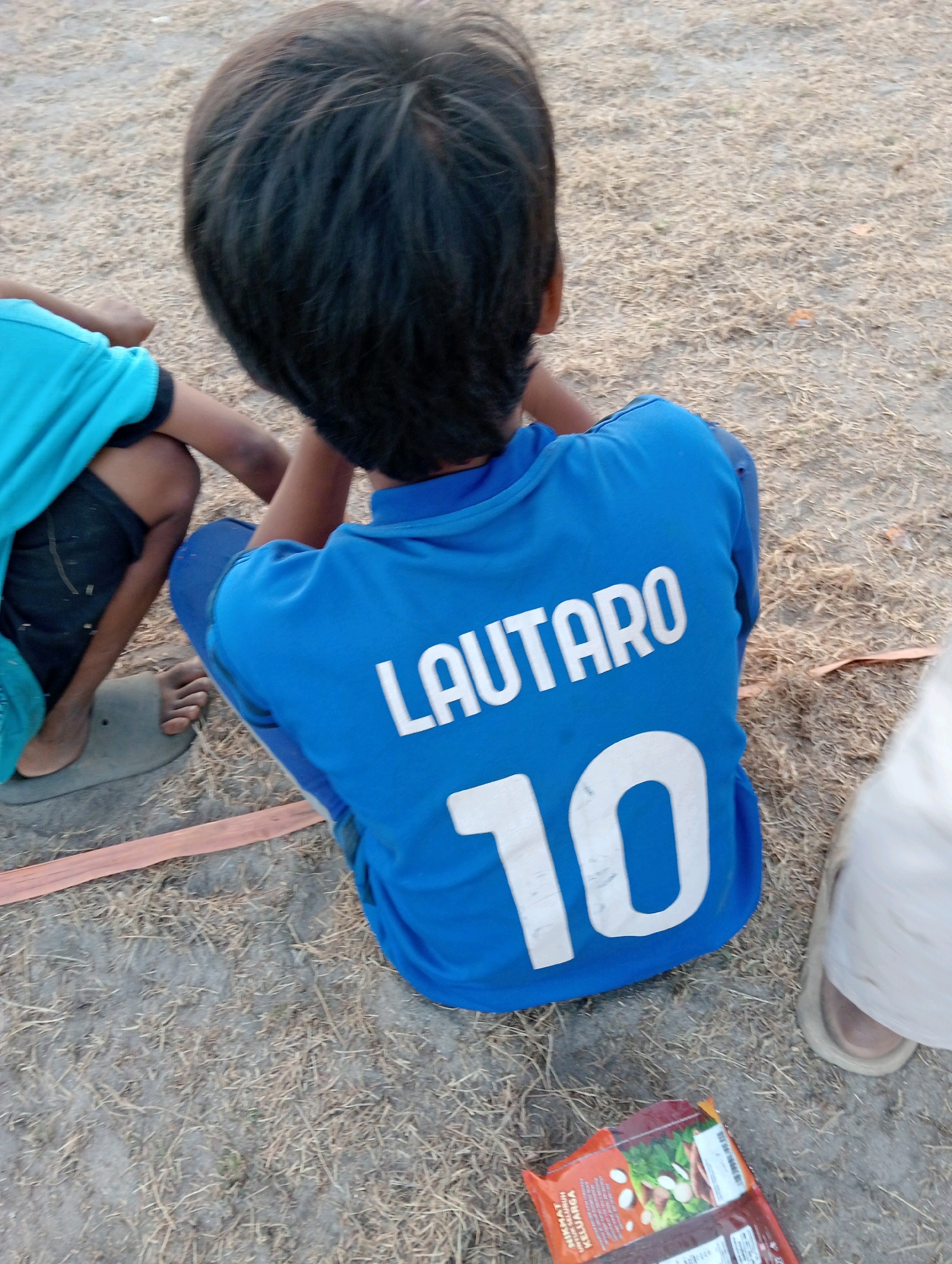 A young boy, viewed from behind and from the right, sits on dry, straw-like grass in Pagerungan Kecil, Indonesia. He wears a bright blue jersey with the name "LAUTARO" and the number "10" printed in white on the back. To his left, another child's legs and torso are visible, clad in a bright turquoise shirt and dark shorts. The ground is a mixture of dry grass and dirt. In the lower right corner of the frame, a person wearing light-colored trousers and a sandal is partially visible. A small, open package of food items with Asian text and images of food lies on the ground near the sandal. A thin, orange strap or ribbon runs horizontally across the lower part of the image.