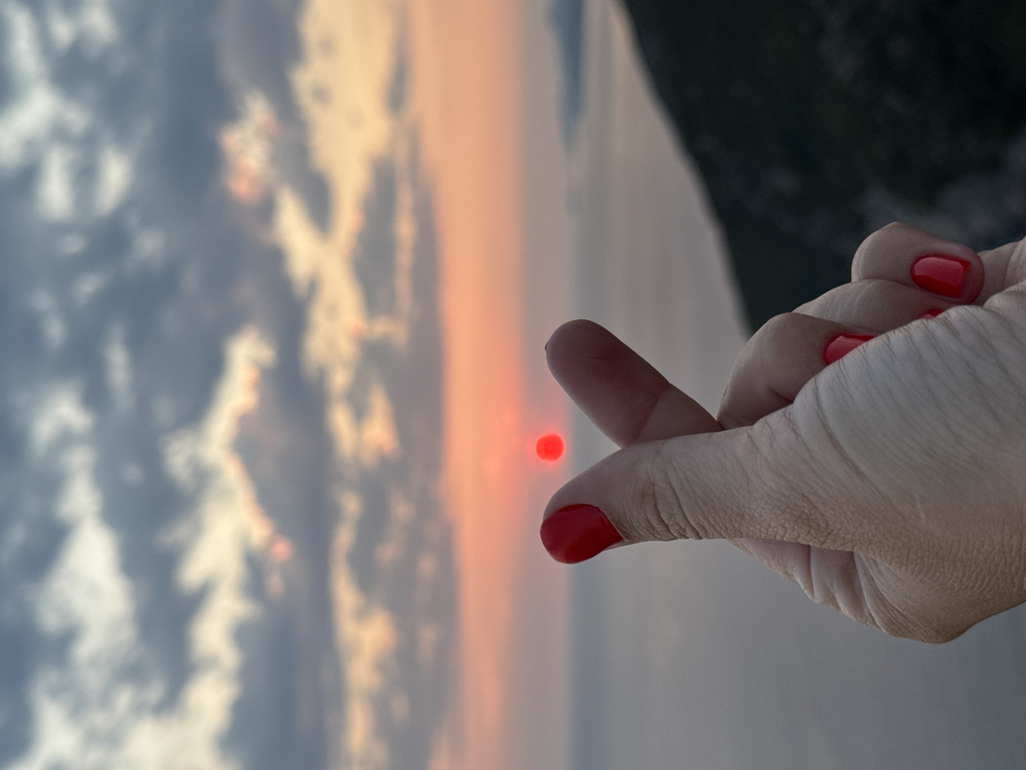 An extreme close-up shot captures a person's hand with bright red nail polish forming the "finger heart" gesture against a blurred background of a sunset over the sea. The sun appears as a small, bright red orb positioned between the thumb and index finger. The sky is filled with a gradient of colors, from soft orange and pink near the horizon to darker gray and blue clouds higher up. The sea is visible in the mid-ground, also blurred, with a dark, hilly coastline on the right side of the frame. The overall impression is a scenic view with a human gesture superimposed. The location is identified as being in the city of Zagora, Montenegro.
