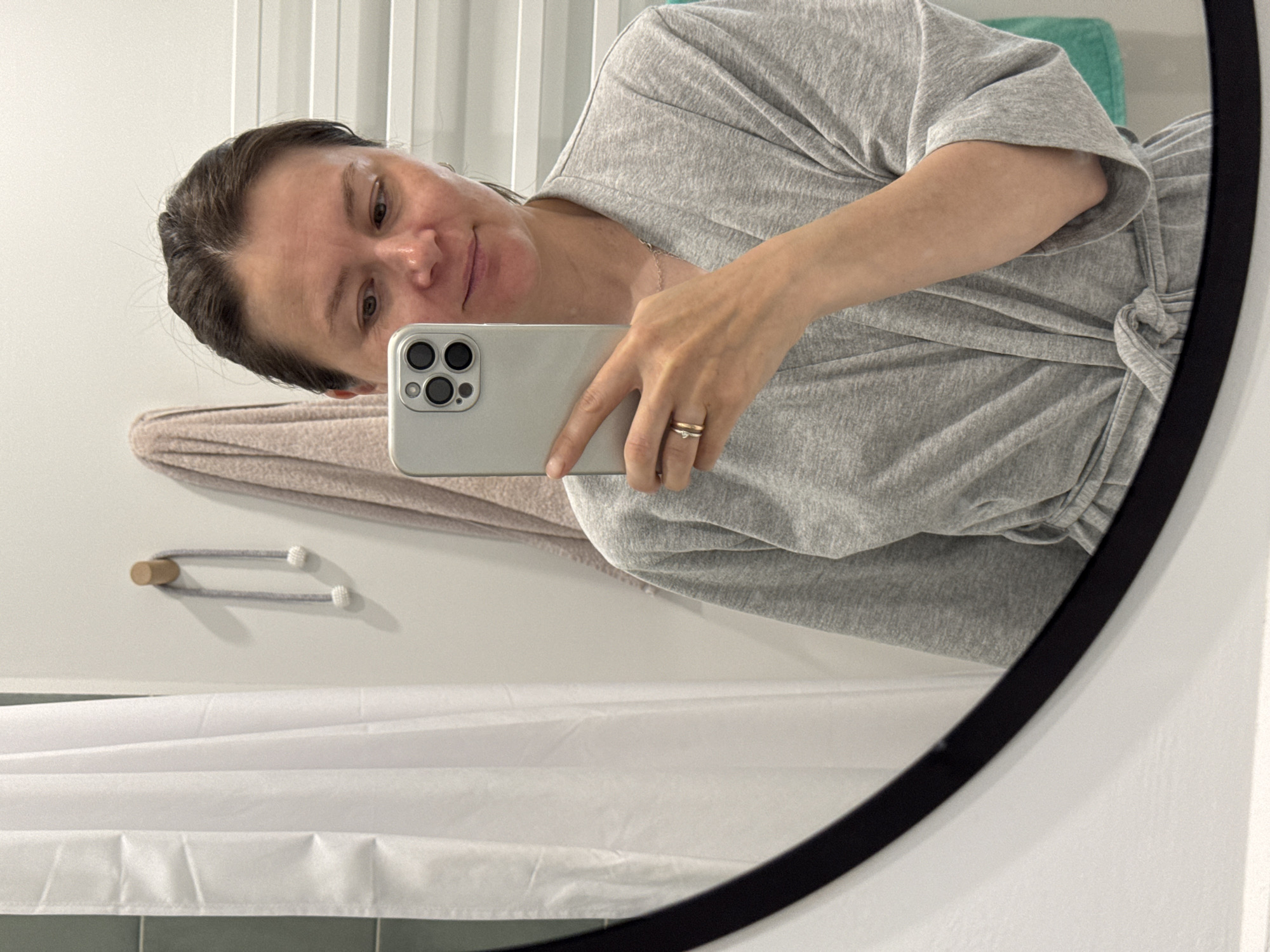 A middle-aged woman is visible in a mirror selfie. She is wearing a grey, belted, wrap-style robe or dress. She is holding a silver smartphone, with her ringed fingers evident. The background reveals a bathroom setting: a white shower curtain, a towel bar, a folded towel in a muted pink color, and part of a teal-colored item (possibly another towel or storage container). There are white vertical blinds behind her. The photograph appears to have been taken indoors in a well-lit room. There is no information that specifies Izhevsk, Russia.