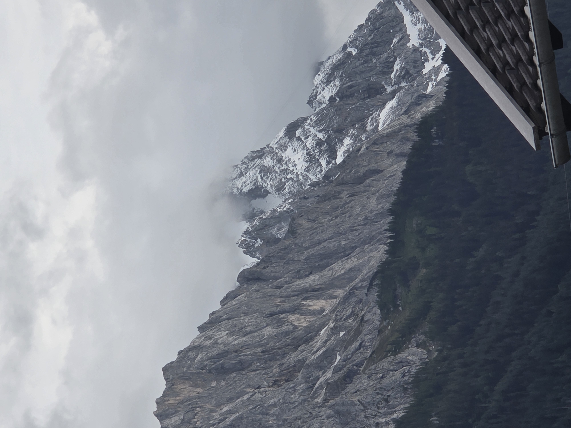 A mountainous landscape in Garmisch-Partenkirchen, Germany is depicted under a cloudy sky. The foreground features a section of a tiled roof with a gutter. In the mid-ground, a dense forest of dark green evergreen trees covers a slope leading up to a large, rugged mountain range. The mountains are characterized by their gray, rocky textures, with patches of snow visible on the higher elevations and peaks. Some of the mountain faces show deep crevices and sharp, jagged edges. The top of the image is dominated by a cloudy sky with varying shades of gray.