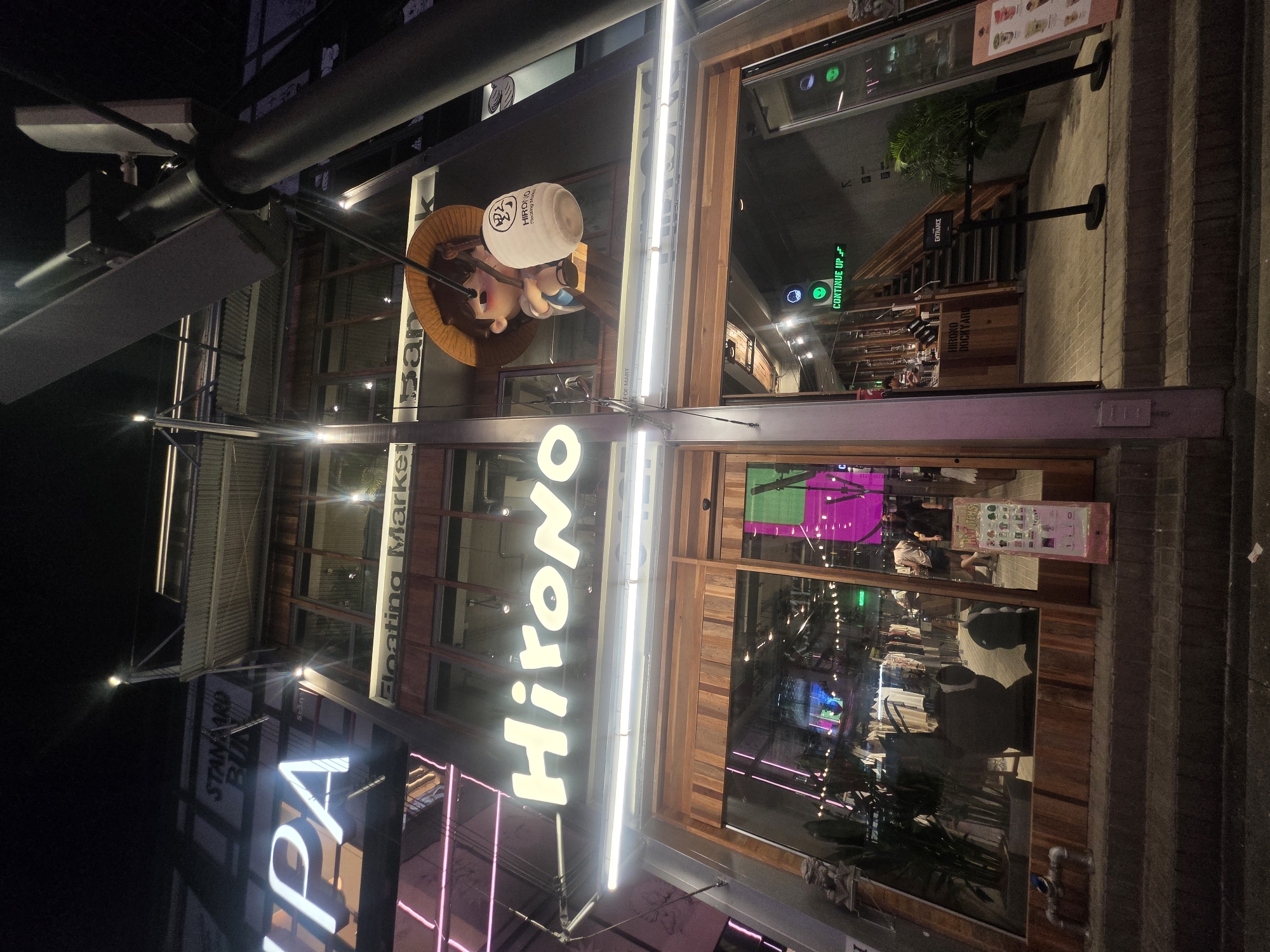 The image shows a storefront in Bangkok, Thailand. The signs read “Standard Spa”, “Floating Market”, “Hirono”, and “Continue Up”. There is a cartoon figure wearing a straw hat and holding a bottle with the words "Hirono Dockyard" on it. Other signs point to “Entrance” and “POP MART”. The store has glass windows showing people inside. The building is lit with fluorescent lights.