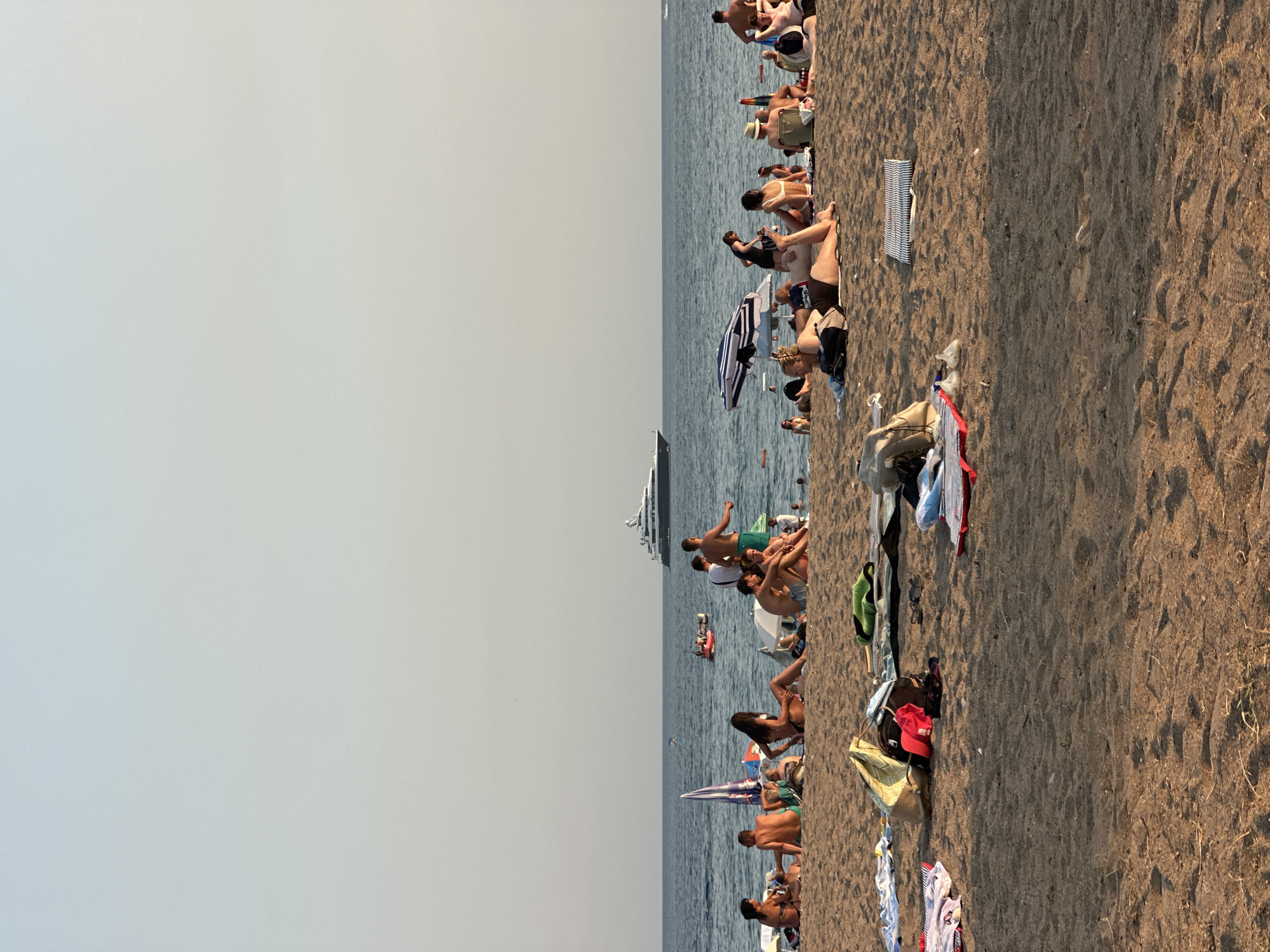 The image depicts a crowded beach scene, likely in Boreti, Montenegro. In the background, a large yacht sails on the calm, blue sea under a clear, pale sky. Closer to shore, people are sunbathing and relaxing on the sand. Some individuals are sitting, while others are lying down, either on towels or directly on the sand. Beach umbrellas are present, providing shade. A few small boats are visible in the water. The foreground is dominated by the sandy beach, with various personal items like bags, towels, and clothing scattered around. The overall atmosphere appears to be one of leisure and summer recreation.