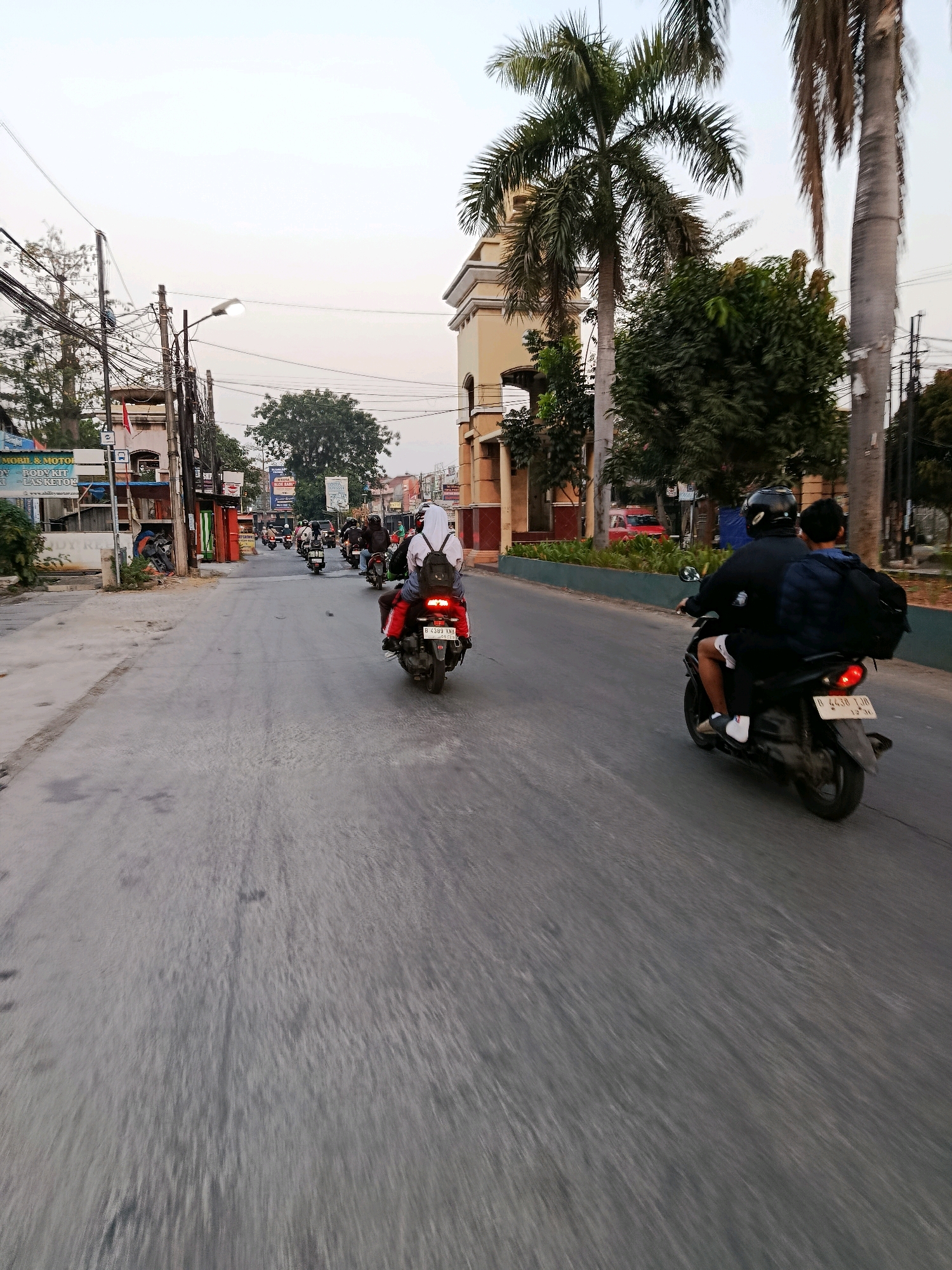 The image shows a street scene in the city of Bekasi, Indonesia, with several motorcycles on a paved road. In the foreground, a black motorcycle carries two passengers, a driver wearing a helmet and a passenger with a blue backpack. The motorcycle's license plate is visible. Further down the road, another motorcycle with a rider wearing a white hijab and a backpack is moving away from the camera. Several other motorcycles are visible in the middle ground, some with passengers. The environment includes buildings on the side of the road, trees, palm trees, and utility poles with many wires. The sky is overcast, suggesting it might be late afternoon or early morning. The overall impression is a typical urban street scene in Indonesia with a focus on motorcycle traffic.