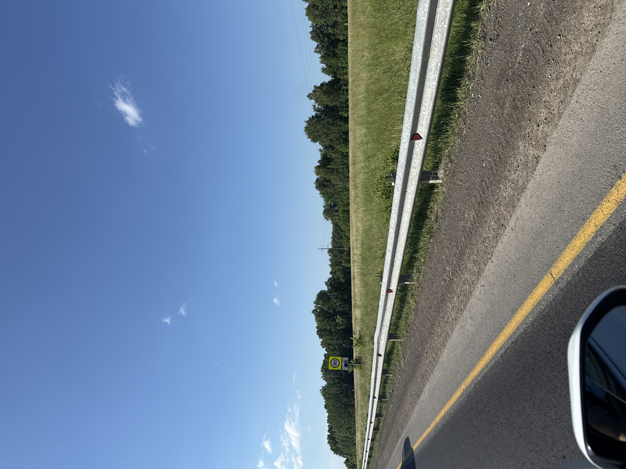The image shows a roadside view on a bright, sunny day. The scene is dominated by a blue sky with a few wispy white clouds. To the right, a metal guardrail runs along the edge of a paved road marked with a yellow line. Beyond the guardrail is a grassy field bordered by a dense line of dark green trees. In the distance, faint power lines are visible against the sky. A speed limit sign indicating "90" is mounted on a pole next to the guardrail. The perspective suggests the photo was taken from inside a moving vehicle. The overall impression is one of a clear, pleasant day, likely in a rural or suburban area, with normal road traffic conditions.