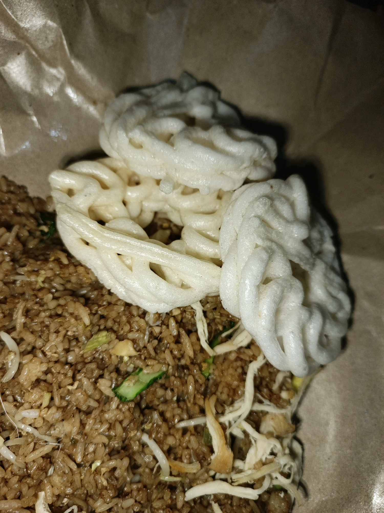 A close-up, overhead shot shows a portion of a brown paper bag containing fried rice topped with two white, coiled crackers and shredded chicken. The fried rice appears to be seasoned and includes visible pieces of vegetables and sprouts. The lighting in the image is dim, creating shadows. The context of Ketegan, Indonesia is not directly depicted in the image itself.