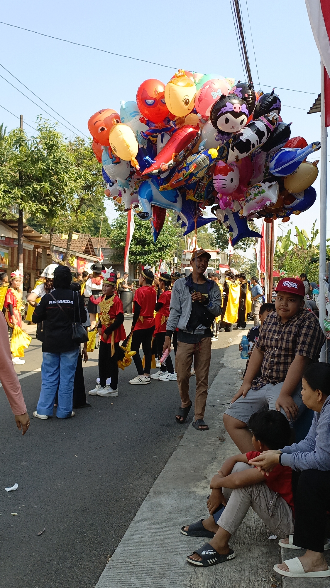 In Krajan Lor, Indonesia, on a sunny day, a vibrant outdoor event unfolds. A man stands by a road holding a large bunch of colorful balloons, some shaped like cartoon characters and animals. In the background, a group of children in traditional red and gold costumes are participating in a parade or performance, holding flags and accessories. Other individuals are gathered, some sitting on a curb and others walking, observing the festivities. The scene is set on a paved road with trees and buildings lining the street, indicating a local community gathering.