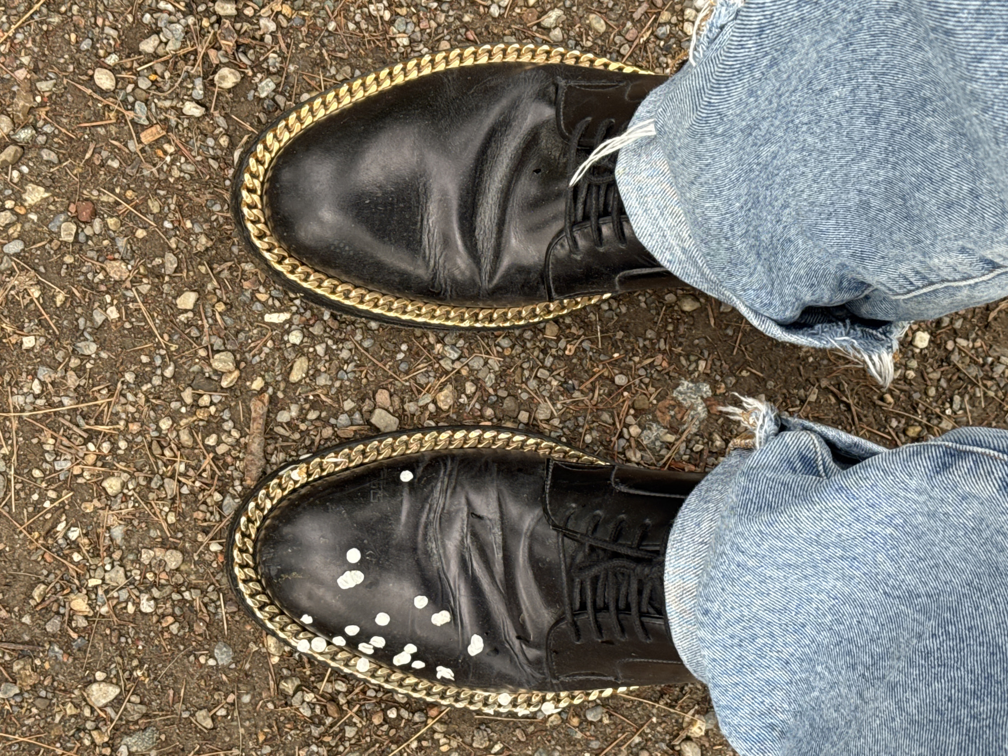 The image shows a downward-angled view of a person's feet standing on gravel and dirt. The person is wearing light blue jeans with frayed edges at the ankles. The jeans are cuffed above the ankles. The person is wearing black leather shoes with a thick gold chain around the edge of the sole. One of the shoes has several white splatters on the front. The background is ground covered with small gravel, dirt, and small twigs. The location is not ascertainable from the image and cannot be definitively said to be Milan, Italy.