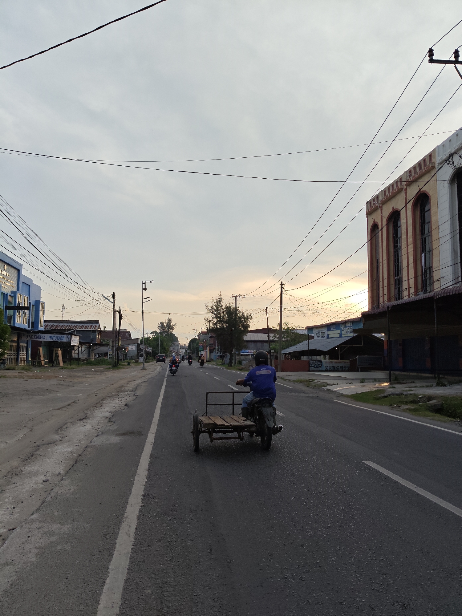 The image shows a street in Lhokseumawe, Indonesia, at dusk. A person on a motorcycle with a trailer is traveling on the right side of the road away from the viewer. Several other motorcycles are further down the road. Buildings line both sides of the street, with power lines overhead. The sky is overcast.