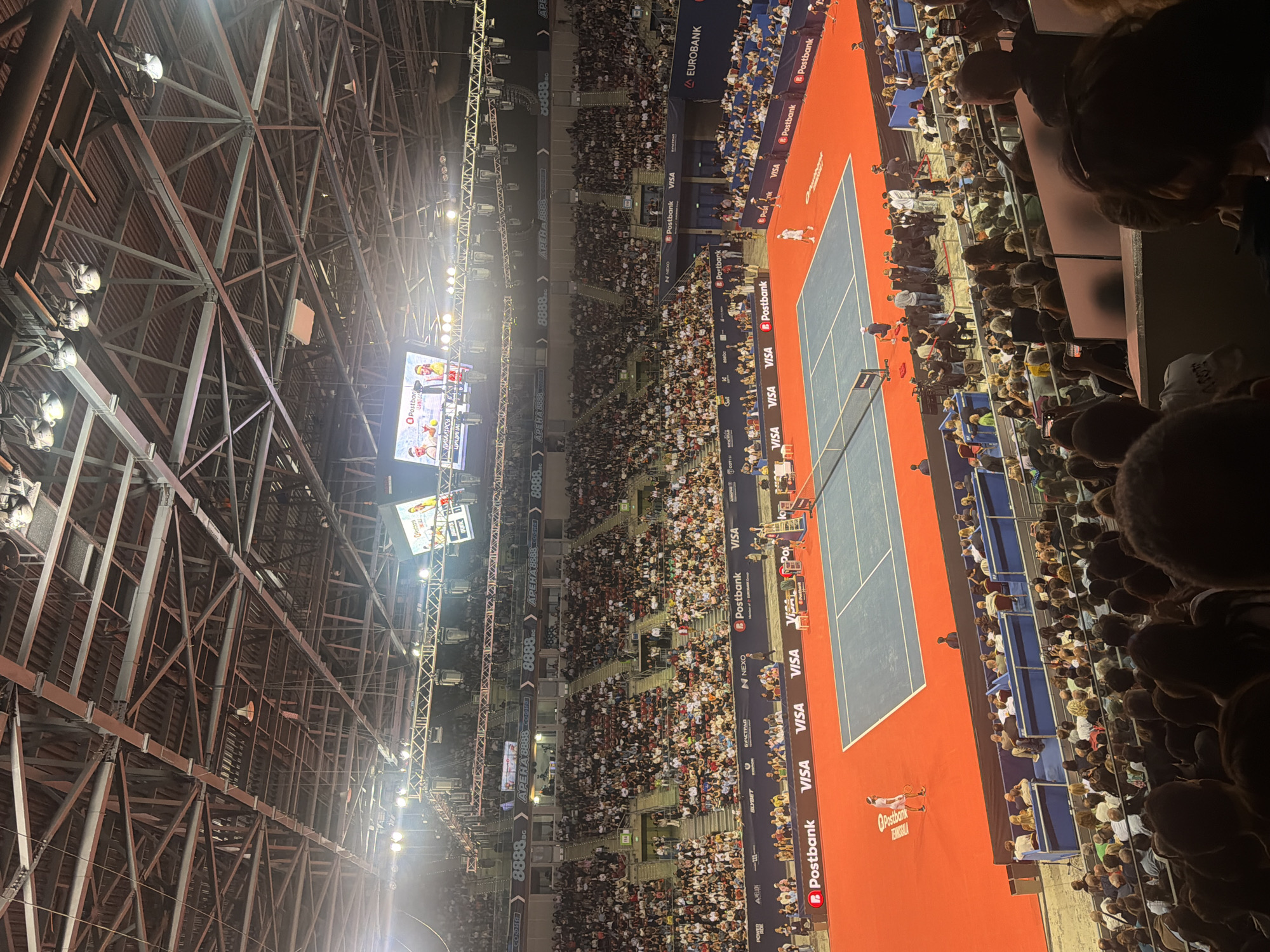 The image shows an indoor tennis match at Arena 8888 in Sofia, Bulgaria. A tennis court with a blue surface and red surround is visible. Two male players are on the court, one in white and the other in blue. A large crowd of spectators is seated in tiered stands surrounding the court. The stadium features a high ceiling with exposed metal beams and lighting rigs. Advertising banners for Postbank and VISA are displayed around the court and on the stadium walls. The overall atmosphere appears to be that of a professional sporting event.