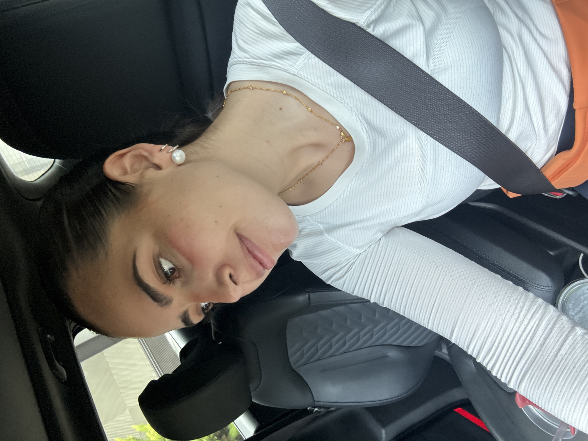 A woman is sitting in a car, wearing a white long-sleeved ribbed shirt and a seatbelt. She has dark hair pulled back, a pearl earring, and a gold necklace. The car interior is black. The location is San Bernardino Tlaxcalancingo, Mexico, as implied by the prompt. The perspective is from a skewed angle, suggesting the image was taken inside a moving vehicle.