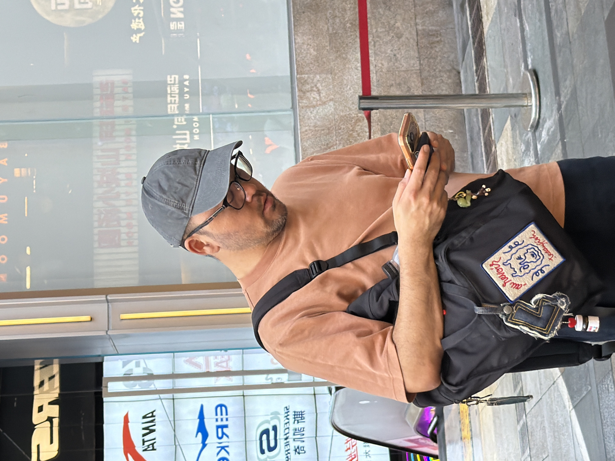 A man wearing a gray baseball cap and a salmon-colored t-shirt is standing on a paved sidewalk in Yuzhong District, China. He is looking down at his gold-colored smartphone, which he holds in his right hand. A black shoulder bag is slung over his left shoulder, and it has several charms attached to it, including a blue denim "L" charm and a patch with a cartoon dog. The background shows a building with large glass windows displaying various signs and graphics, some of which appear to be in Chinese. A red rope barrier is visible in the mid-ground, and a silver metal pole stands to the right of the man. The overall environment suggests an urban street scene.