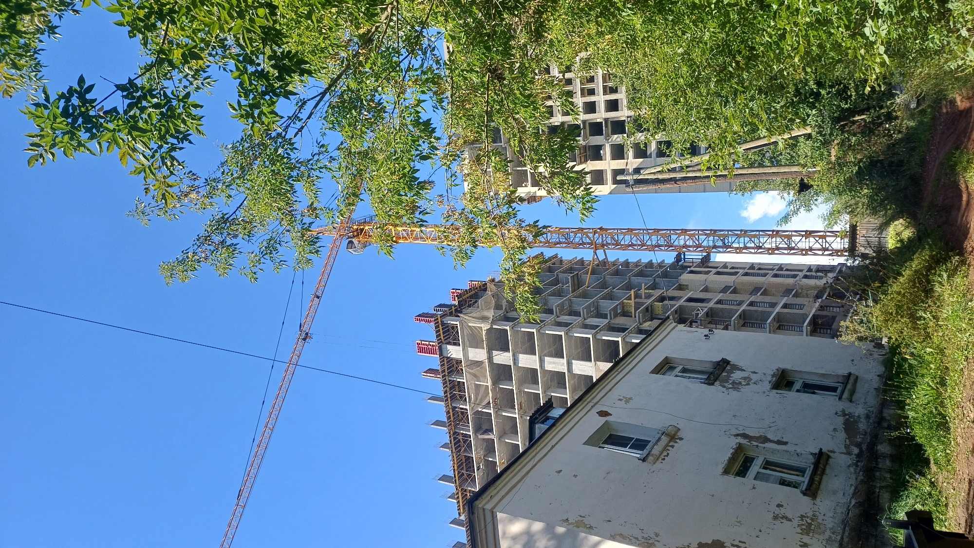 An outdoor, ground-level, eye-level shot captures a construction site under a clear blue sky in Izhevsk, Russia. In the foreground to the left is an old, dilapidated white building with peeling paint and three visible windows. To the right and center are two unfinished high-rise apartment buildings, characterized by their exposed concrete skeletons and grid-like structures. A tall yellow construction crane stands prominently between these buildings, with its boom extending horizontally across the upper portion of the frame. Lush green trees and foliage partially obscure the view of the buildings on the right. The ground is covered in grass and a dirt path. The scene suggests ongoing urban development and contrasts old structures with new construction.
