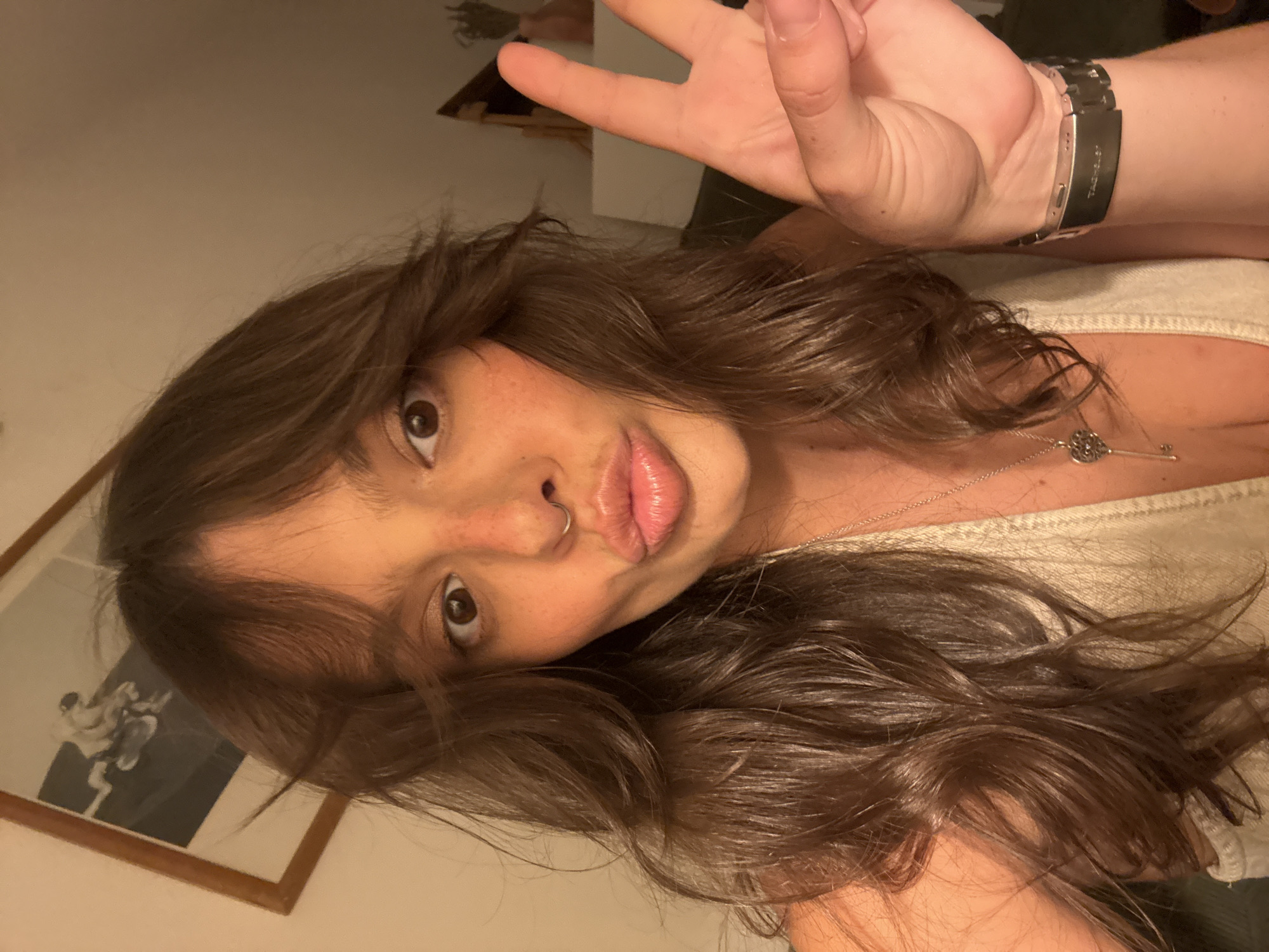 A young woman with long, dark brown hair and fair skin is posing for a selfie. She is holding up a peace sign with her right hand. She has brown eyes, a nose ring, and freckles. She is wearing a light-colored top and a silver necklace with a key pendant. On her left wrist, she wears a silver wristwatch. A framed picture hangs on the wall behind her. The location in Tel-Aviv, Israel is unknown.