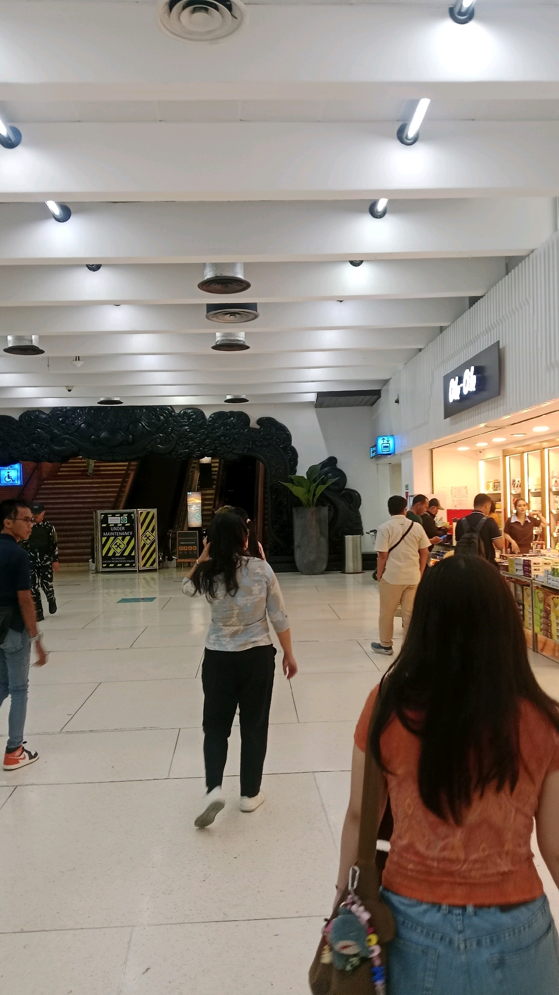 The image depicts an indoor scene, likely a public space such as an airport terminal or a shopping mall in Tangerang, Indonesia. The ceiling features a modern design with multiple white beams, recessed lighting fixtures, and circular air vents. In the background, there's a prominent dark, ornate structure resembling a large carved archway or entrance with stairs behind it. To the right, a retail store with bright interior lighting and merchandise displays is visible. Several individuals are present in the scene, walking and interacting. One person in the foreground is wearing a dark blue t-shirt and jeans, while another, further back and facing away, wears a light-colored floral top and black pants. Other individuals are seen walking in the background and near the store. The floor is tiled with a light-colored material. The overall impression is that of a busy commercial or transit area.