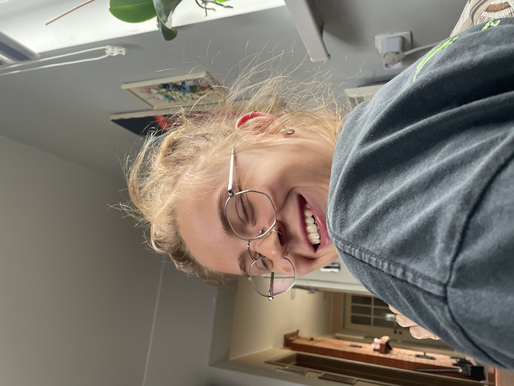 A woman with light blonde hair and glasses is smiling at the camera. She is wearing a dark grey shirt. The photo appears to be taken indoors with a view into another room featuring what looks like a wooden countertop or desk and window. There is no evidence to suggest the location is Sestroretsk, Russia.