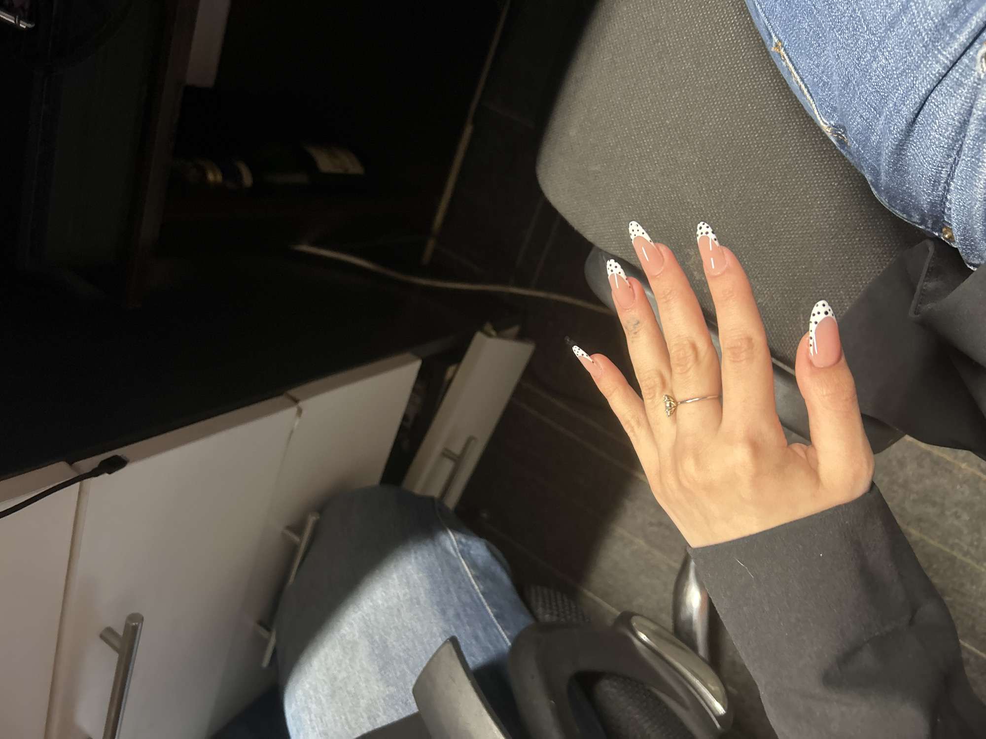 A person's hand with French tip nails adorned with black polka dots is resting on their lap. A ring with a floral design is visible on the ring finger. The person is wearing a black sweater and blue jeans. The background suggests an indoor setting, possibly an office or a room with furniture. There are no identifiable landmarks or specific features that would indicate the location as Tbilisi, Georgia.