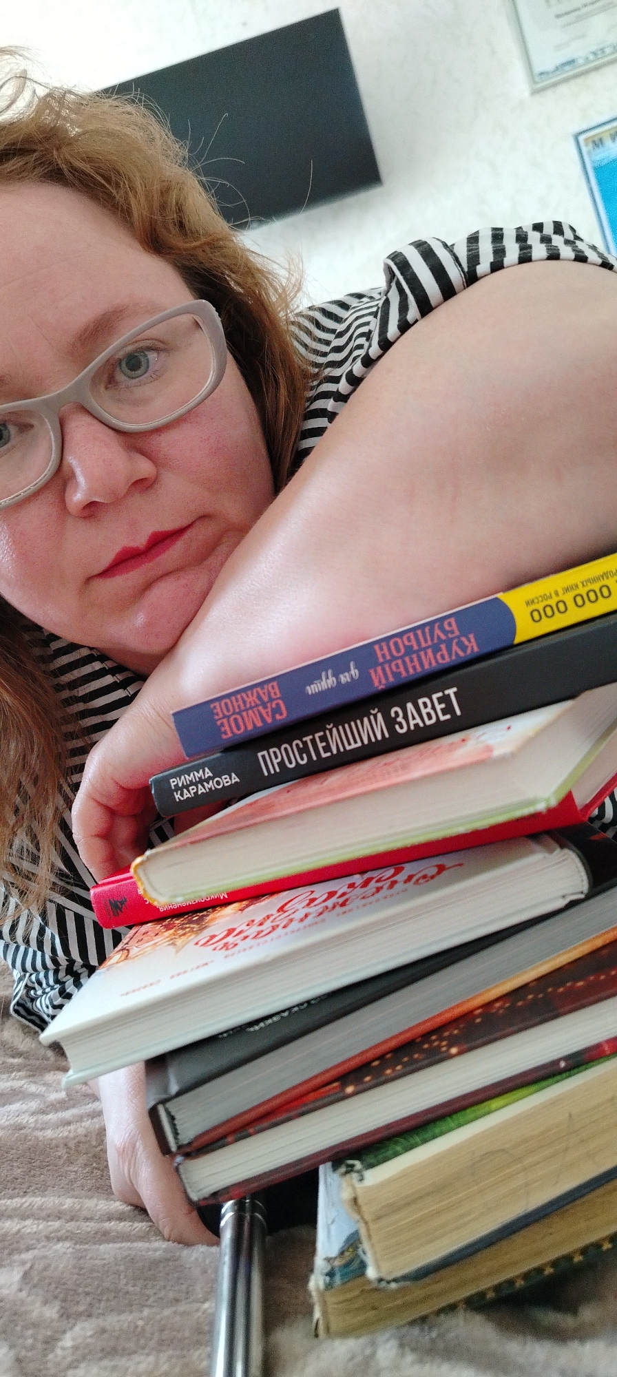 A woman wearing eyeglasses and a striped shirt is resting her arm on a stack of books. The book covers in the stack include Russian titles: "CAMOE ВАЖНОЕ", "ПРОСТЕЙШИЙ ЗАВЕТ", and "БУЛЬОН КУРИНЫЙ для души". The text on the yellow book cover also indicates "РОДАННЫХ КНИГ В РОССИИ". The books and the woman are indoors. There is no indication that this image was taken in the city of Chaikovsky, Russia.