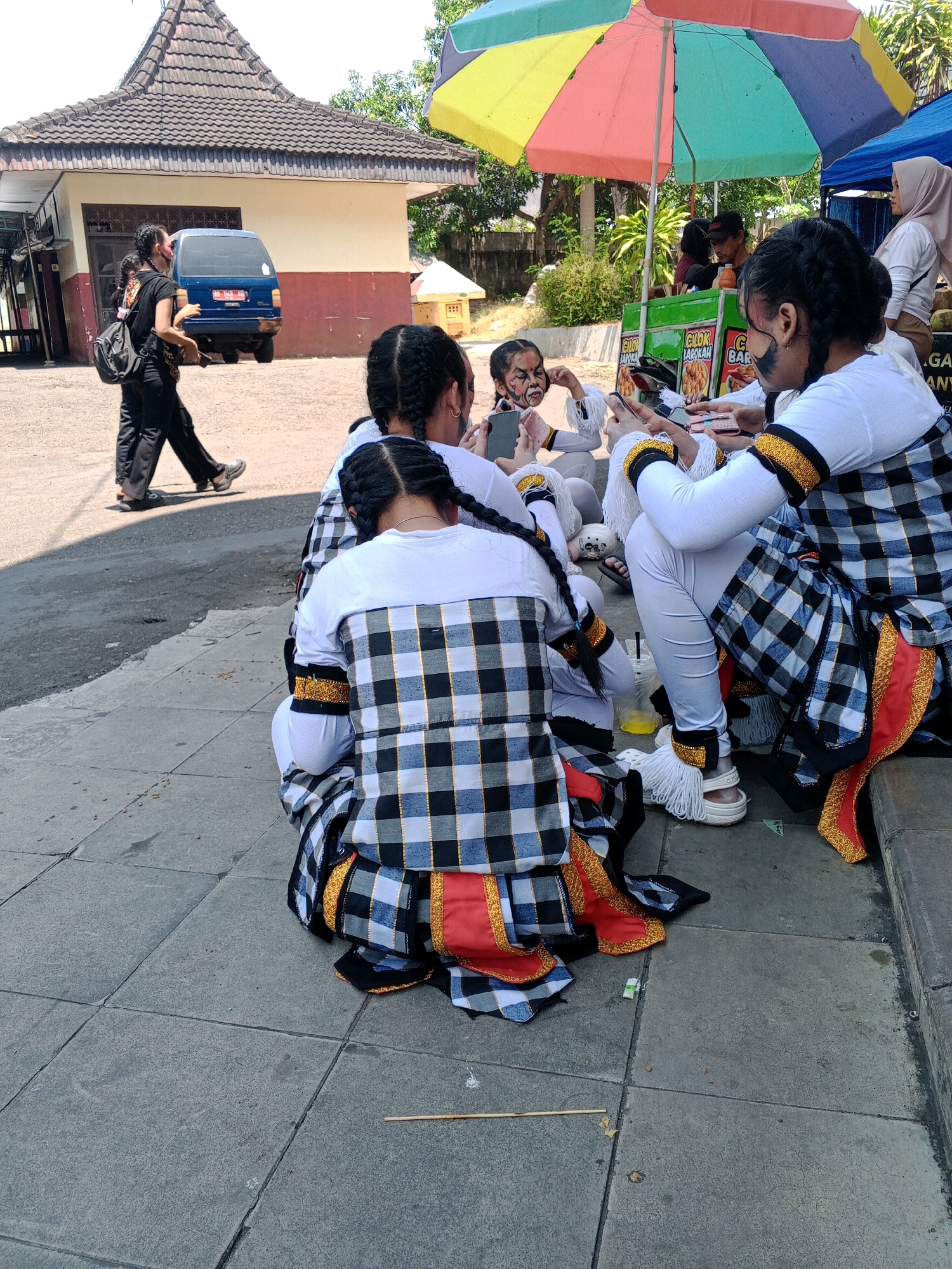 In Wuryorejo, Indonesia, a group of young people dressed in traditional checkered outfits are gathered on a sidewalk. Some are seated, while others are standing. The individuals have distinctive face paint, with one person appearing to have a white face with dark markings and another with dark eyebrows and cheek markings.
Several are looking at their mobile phones, indicating modern technology is present amidst the traditional attire. In the background, a blue van is parked near a building with a tiled roof. A colorful umbrella provides shade for a food cart, with signage indicating it sells "Cilok Barokah" (a type of Indonesian snack). The environment appears to be an outdoor, possibly public, space with a paved sidewalk and some greenery. The overall scene depicts a blend of traditional culture and contemporary life.