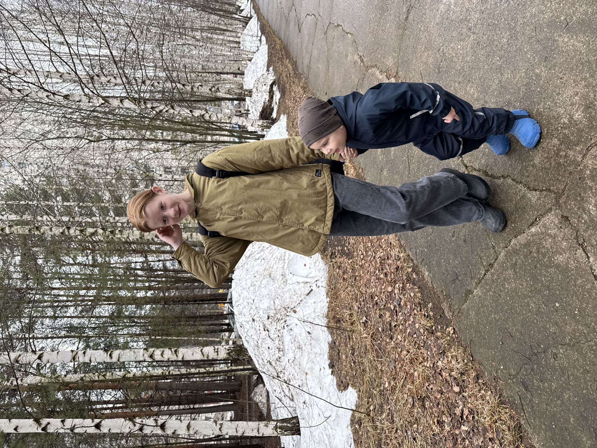 Two individuals are captured outdoors amidst a sparse, late-winter scene. The older person, a teenager, is positioned to the left, donning a khaki jacket, dark jeans, and a backpack. They are making a playful gesture near their ear. To the right is a younger child, clad in a dark blue jacket and blue boots. The child is bent over slightly, hands positioned near the older person's torso.
The location appears to be a park or wooded area, based on the presence of birch trees lacking leaves. Patches of snow are visible on the ground near the trees. A concrete walkway or road runs horizontally through the frame. The ground is covered with fallen leaves and dead vegetation. This scene is most likely somewhere in or around the city of Chaikovsky, Russia.