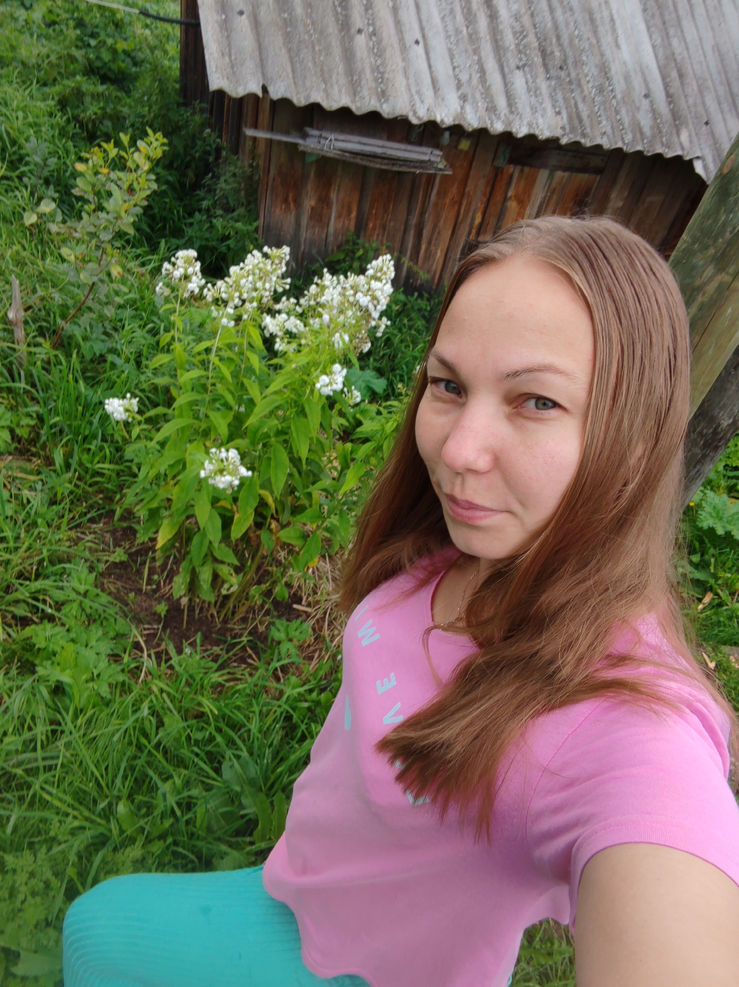 A young woman with shoulder-length brown hair is taking a selfie outdoors. She is wearing a pink t-shirt with light blue lettering and teal leggings. The background shows a wooden shed with a corrugated roof, surrounded by lush green vegetation and some white flowers. The image appears to be taken in an outdoor setting, possibly a rural area or a yard in Chukhlem, Russia.