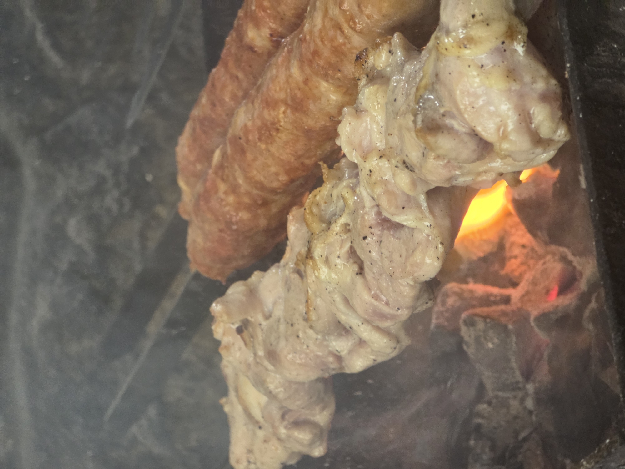 The image shows food cooking over an open flame. The main subjects are several sausages and pieces of chicken, skewered and arranged vertically. The sausages are elongated and have a reddish-brown, slightly textured casing, suggesting they are grilled or roasted. The chicken pieces are lighter in color, with visible seasoning, and appear to be partially cooked, with some parts showing browning.
The scene is set in an outdoor cooking environment, likely a barbecue or grill. Embers and flames are visible beneath the food, indicating the heat source. Smoke is also present, contributing to the atmosphere of cooking. The background is dark and somewhat indistinct, possibly suggesting a cooking pit or a grill enclosure.
The activity taking place is the grilling or roasting of meat. The food is positioned close to the heat source, implying it is in the process of being cooked. There are no people visible in the frame, so the focus is solely on the food and the cooking process.
Notable details include the texture of the sausages and the way the chicken is seasoned. The bright orange and red hues of the flames contrast with the muted tones of the meat and the dark background. The presence of smoke adds a sensory element to the visual. No visible text is present in the image. The context provided is Pidhorodne, Ukraine, which suggests this is likely an outdoor meal preparation in that location.
