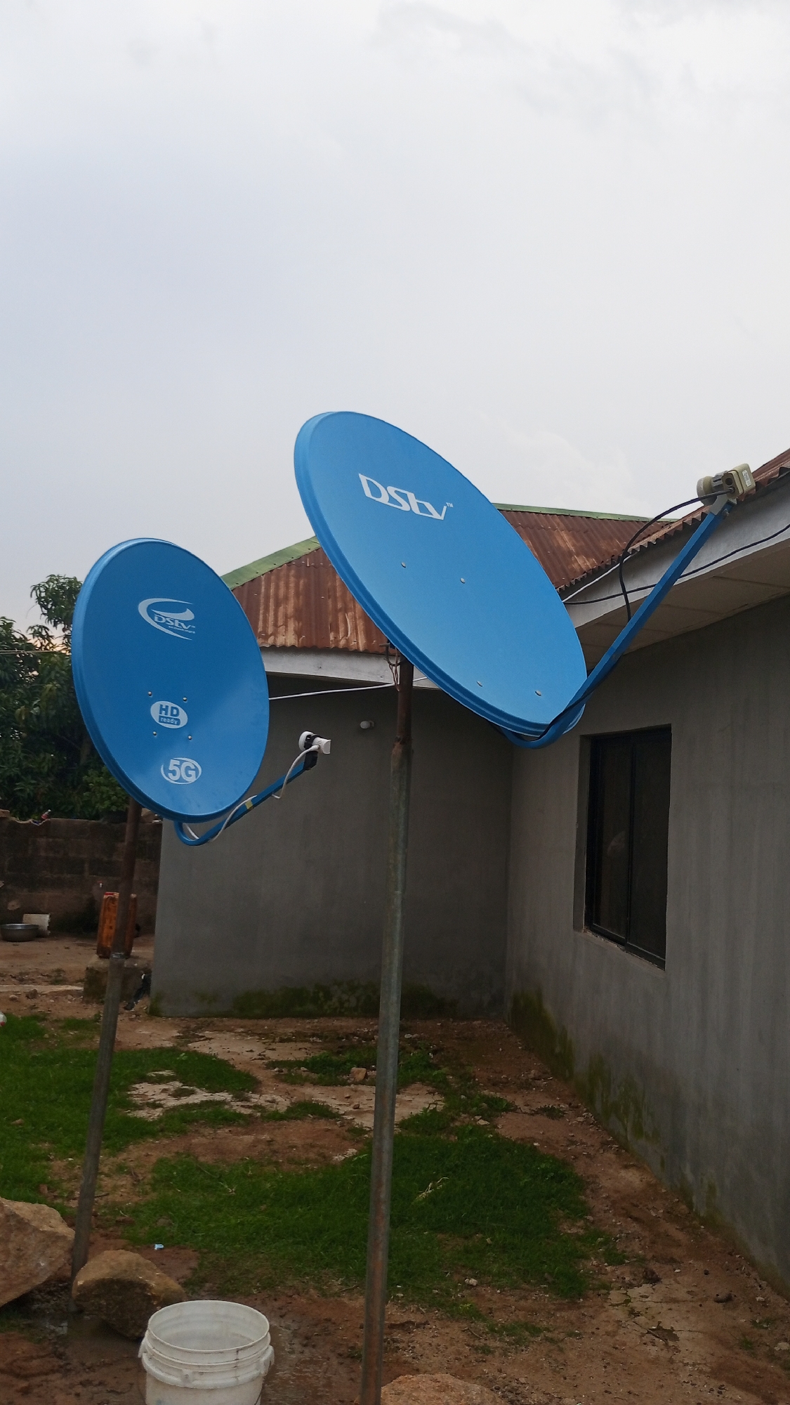 The image depicts an exterior view of a building with two blue DStv satellite dishes. One dish is mounted on a pole in the foreground, and the other is attached to the side of the building via an arm. The building has a corrugated metal roof and a grey concrete exterior wall with a dark-framed window. The ground is a mix of dirt, patches of grass, and stones. A white bucket is visible on the ground in the lower left corner. The sky is overcast. The scene is located in Barikin Sabon Gidan Kanat, Nigeria.