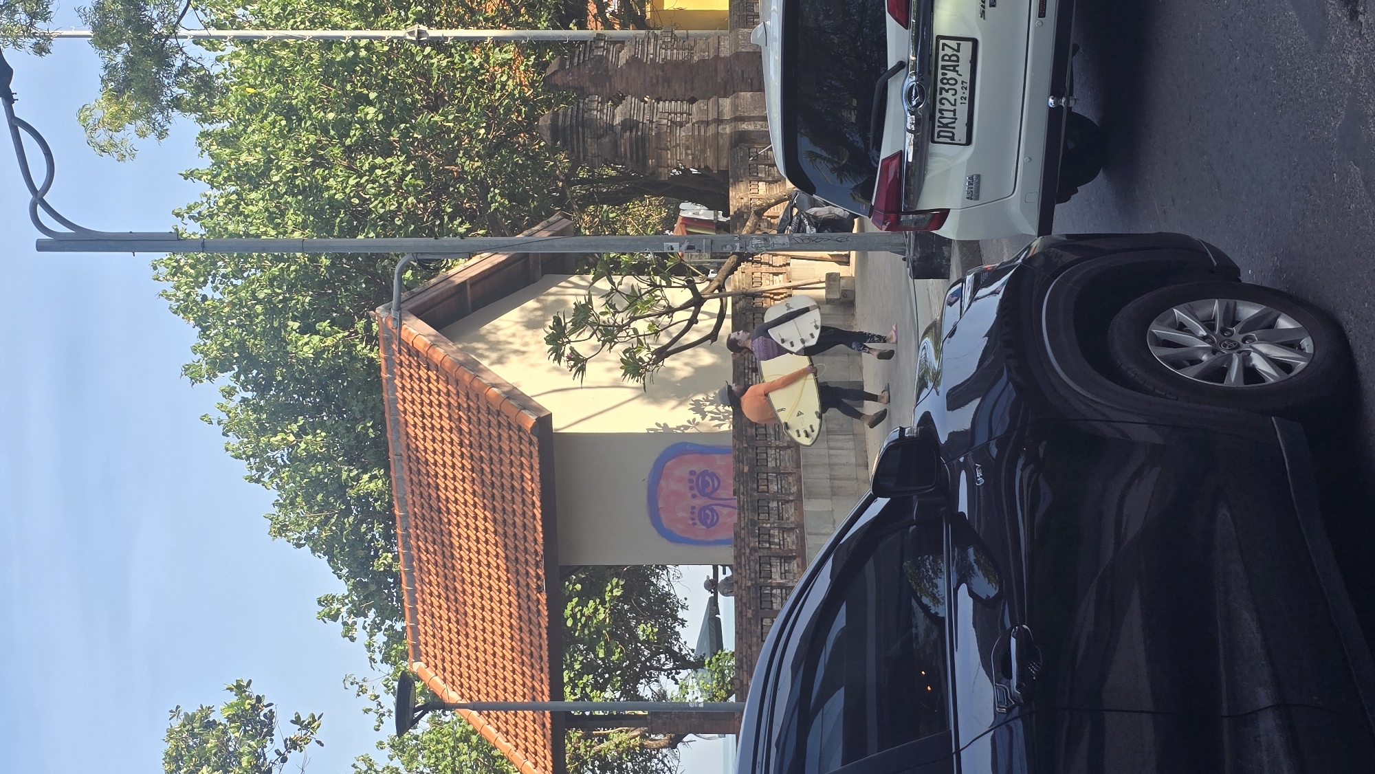The image depicts a street scene in Kuta, Indonesia. Two people are walking on a sidewalk, each carrying a surfboard. A building with an orange tiled roof and a mural is behind them. Two cars, one white and one dark, are parked on the street next to the sidewalk. Trees and a lamppost are visible in the background. The license plate of the white car reads "DK1238ABZ". The sky is blue.