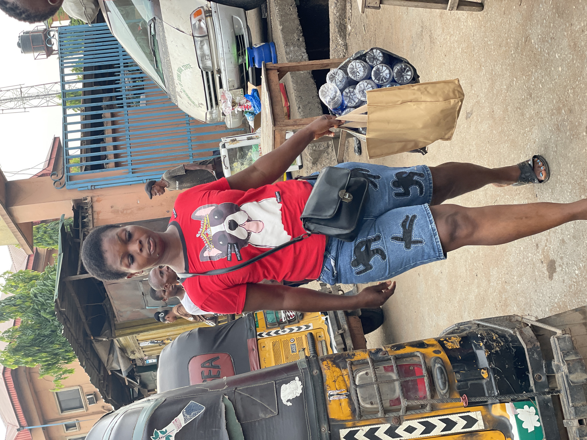 This outdoor, daylight scene captures a bustling street or market area, likely in Onitsha, Nigeria, given the visible tricycle (Keke). The central focus is a young person with dark skin and short dark hair, standing upright and looking towards the camera. They are wearing a red t-shirt featuring a graphic of a bulldog with sunglasses and the text "DOLCE GABBANA" on its chest. They also wear blue denim shorts with black outline patterns, black sandals, and a black shoulder bag. Their toes have light blue nail polish. They are holding a light brown paper bag near a makeshift wooden stand filled with plastic bottles of water, suggesting a purchase or sale.
In the background, to the left, a white commercial van or bus is parked, partially obscured by a blue metal gate or fence. Text partially visible on the van includes "GOVERNMENT" and "STATE". Further back, a water tank is mounted on a structure. To the right and behind the main subject, a yellow and black Keke (tricycle) is prominent, displaying "AREA" on its seat cover and "AC" on a yellow vent. Various stickers and patterns adorn the Keke. Several other individuals are visible in the mid-ground, mostly partially obscured by the main subject or vehicles; one person wears a dark shirt and cap. The ground is a light-colored paved or concrete surface. The lighting is bright, indicating daytime, possibly overcast or late morning/early afternoon.