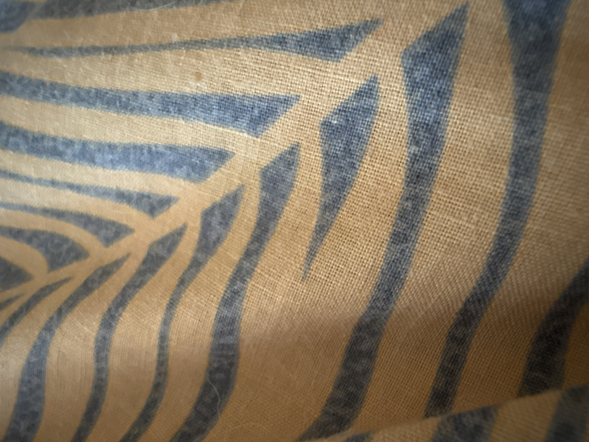 The image shows a close-up of a textured fabric with a striped pattern. The stripes are alternating between a warm, light brown color and a darker, grayish-blue color. The fabric appears to be woven, with visible threads creating a slightly rough texture. The pattern is abstract and organic, with the stripes varying in width and curvature. The lighting highlights the texture of the fabric, casting subtle shadows that give it a three-dimensional quality. There are no discernible people, objects, actions, or interactions visible in the image. The environment is not discernible as there is no background present. The image appears to be a photograph of a textile, possibly a scarf, blanket, or piece of clothing. There is no indication of any location in the city of Okhtyrka, Ukraine.