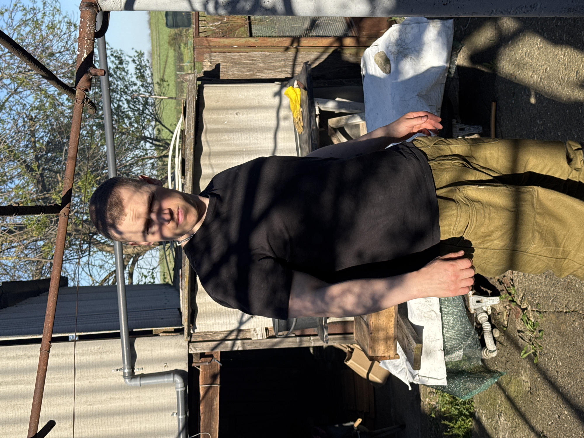 The image depicts a person with short brown hair wearing a black t-shirt and olive green pants, standing outdoors near a corrugated metal building. Overhead are pipes and tree branches. Shadows are prominent, cast by sunlight. A white tarp and some other objects are on the ground. The location is likely Goncharka, Russia, given the prompt.