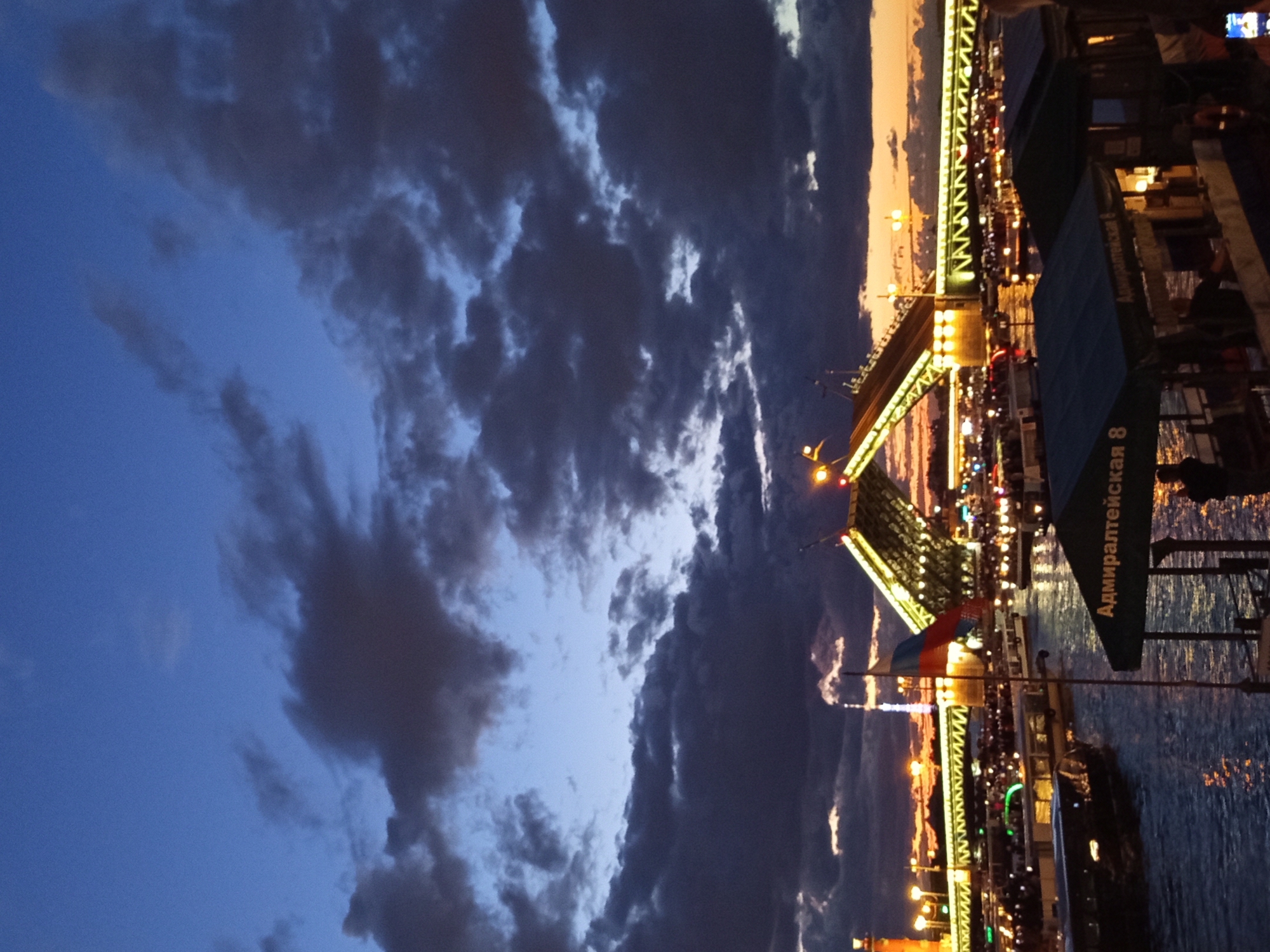 The image depicts a twilight scene of the Palace Bridge in Saint Petersburg, Russia. The bridge is raised, revealing its central span elevated over the Neva River. Numerous boats are visible on the water, some docked and others navigating the river. Several boats appear to be passenger ferries, indicated by the presence of people on deck and the illuminated awnings. One awning has Cyrillic text that reads "Адмиралтейская 8" (Admiralteyskaya 8). The sky is filled with dramatic, dark clouds against a deep blue and orange sunset, creating a striking contrast. Lights from the city and the bridge illuminate the water and the surrounding infrastructure, reflecting on the river's surface. A Russian flag is prominently displayed on a flagpole in the foreground. The overall atmosphere suggests a popular tourist spot during the evening, likely a period of white nights in Saint Petersburg.